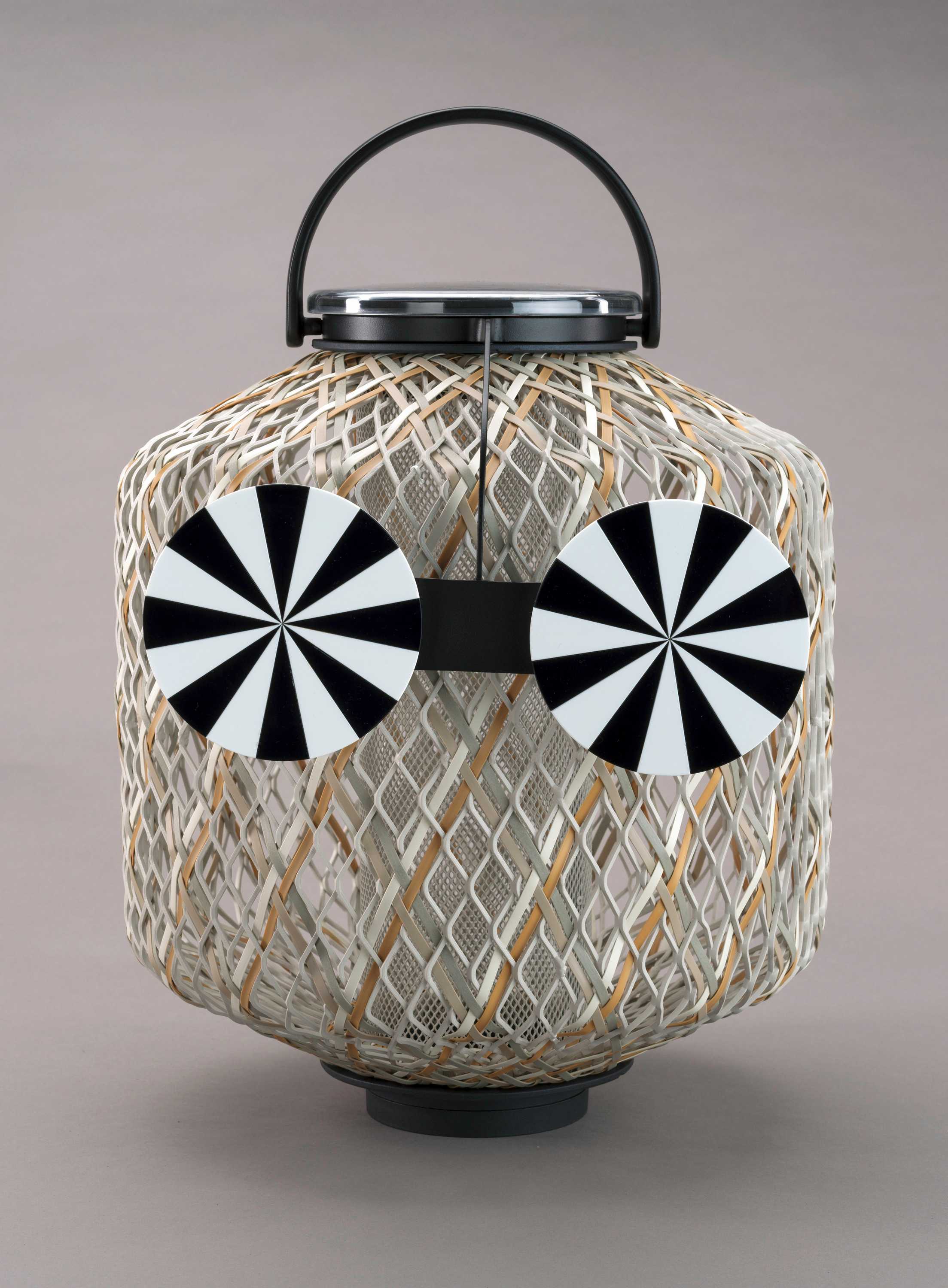 Lantern  with white-coated aluminum frame with interwoven fibers.  It sits on a black base and has a curved black handle on top.