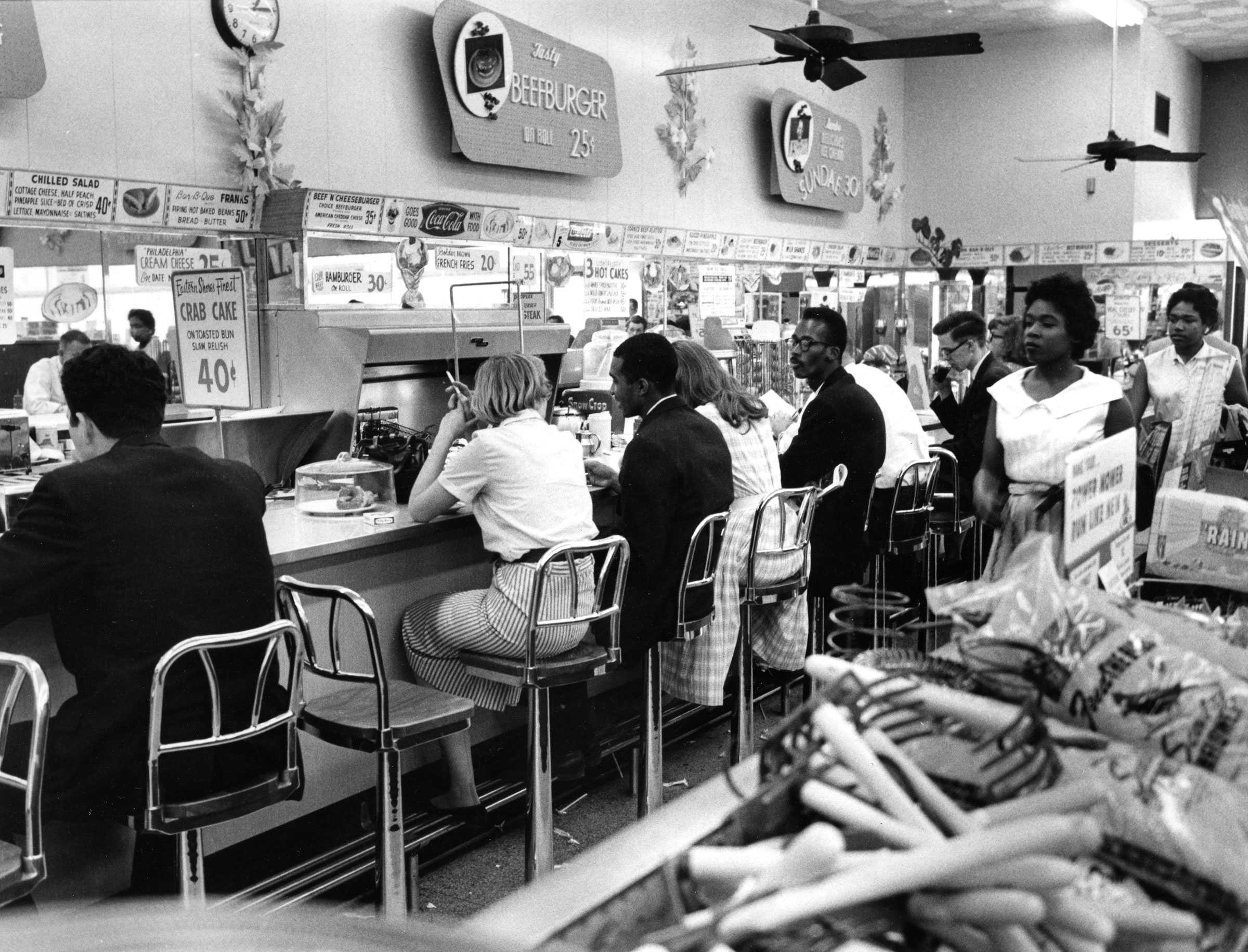 A black and white photograph of a group of black and white people sit at a counter at People's Drugstore Arlington, Virginia.