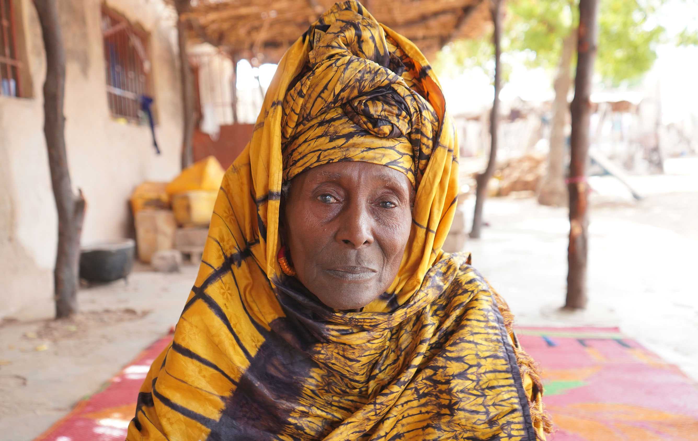 An older woman wearing a black and yellow headwrap and covering. She is look directly at the camera with a slight smile.