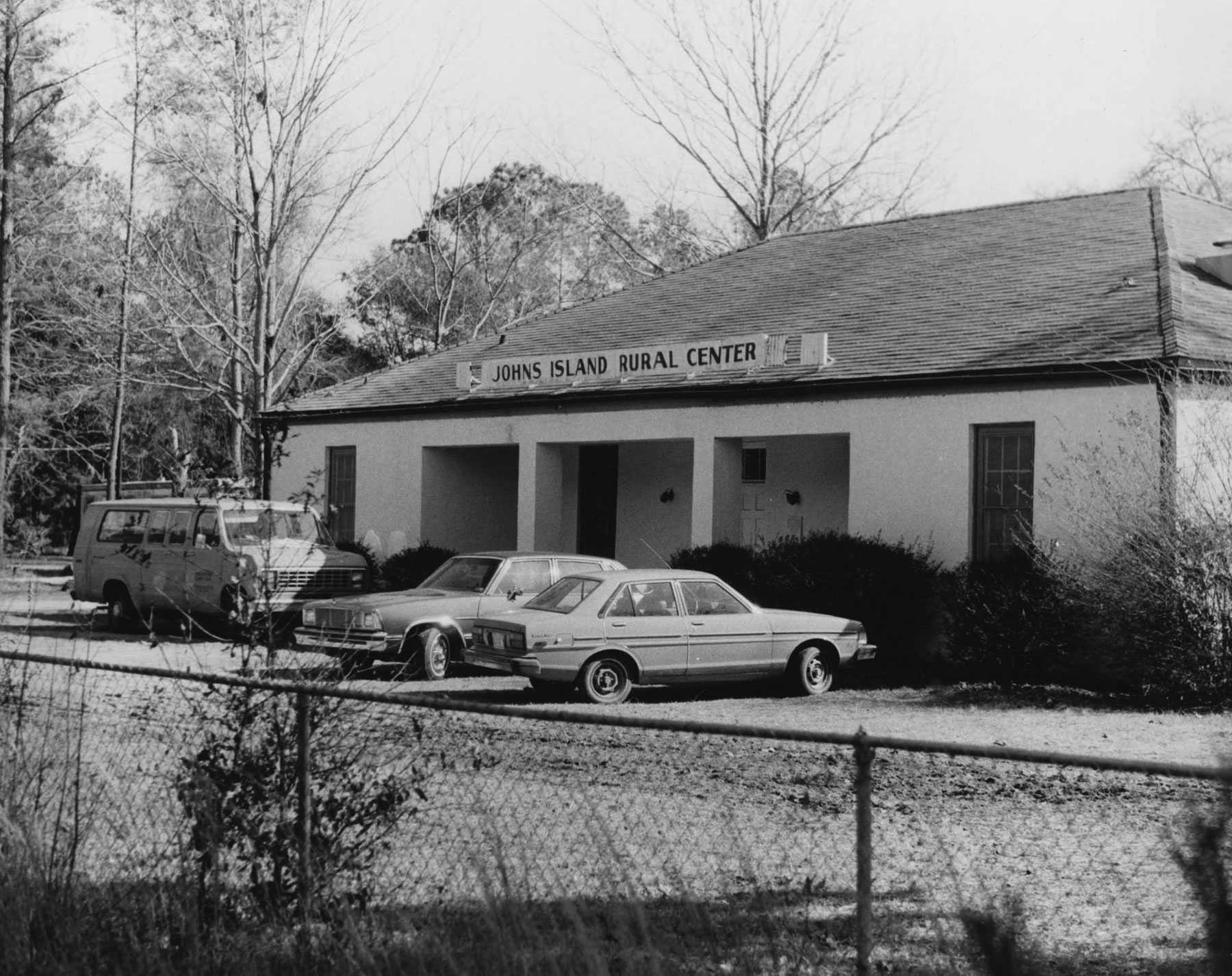 A black and white photo of the outside of the Johns Island Rural Center. Two cars and a van are parked in front.