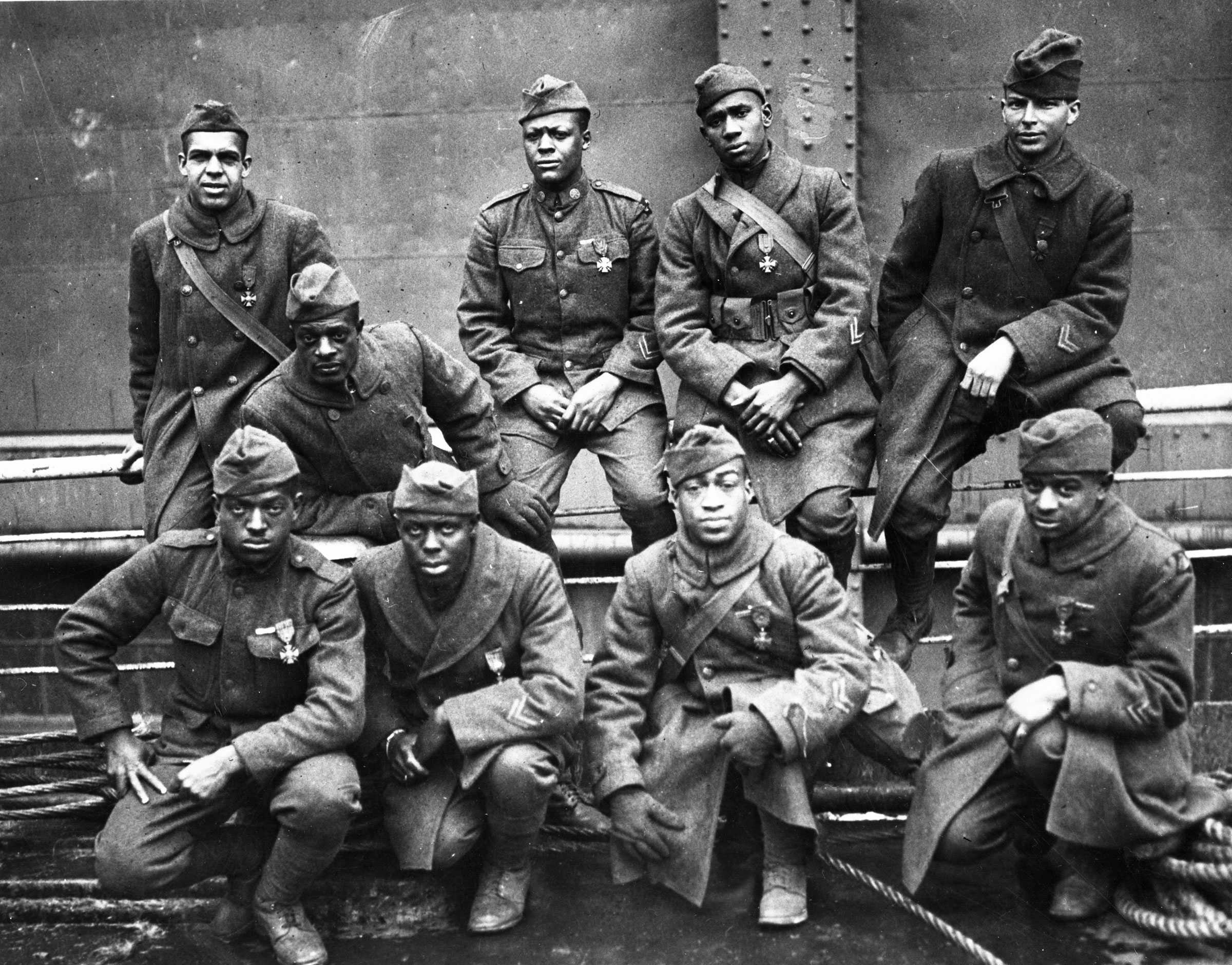 Black and white group portrait of nine men dressed in military uniform standing before a ship.  They all have a "medal pinned to their uniforms that is a cross.