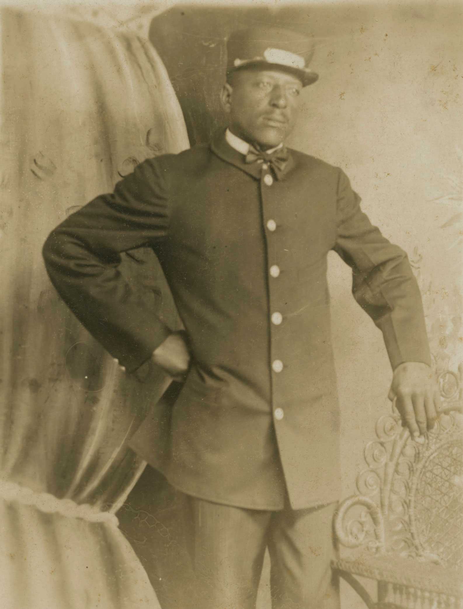 Black-and-white postcard of Pullman Porter T.R. Joseph in uniform, posing in a studio with a theater backdrop.