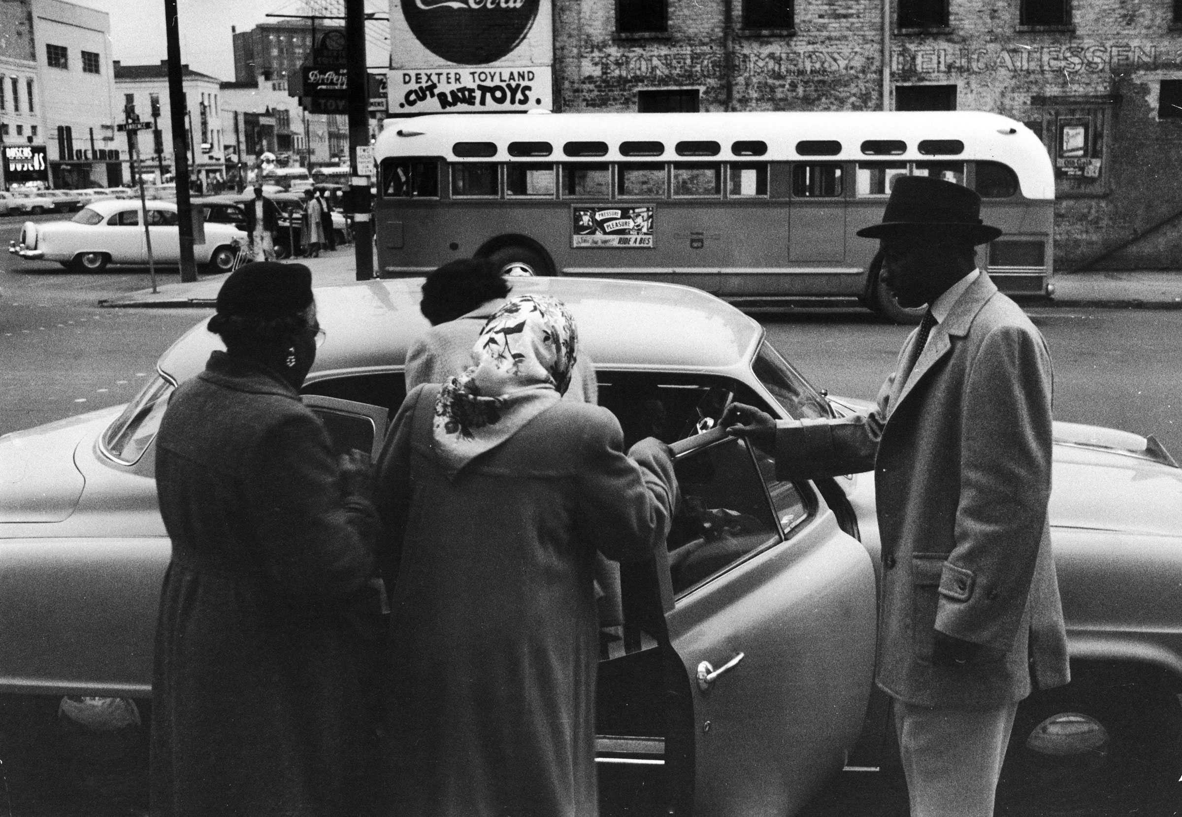 A black and white photograph of older ladies getting into a car. A gentleman holds the doors open as they climb into the back seat.