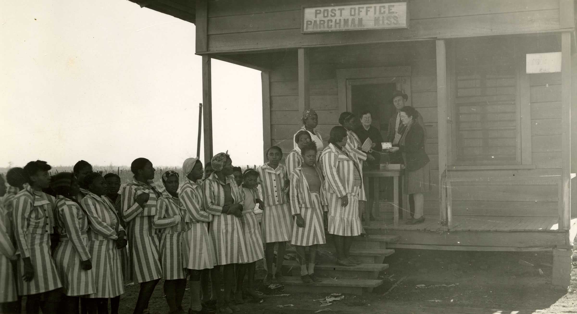 Black and white photograph of Black women lined up in front of a small wooden house with signage "POST OFFICE / PARCHEMAN, MISS.  The women are dressed in striped prison clothing.  There is a table set up at a doorway where two White women and a White man distribute items to the women.