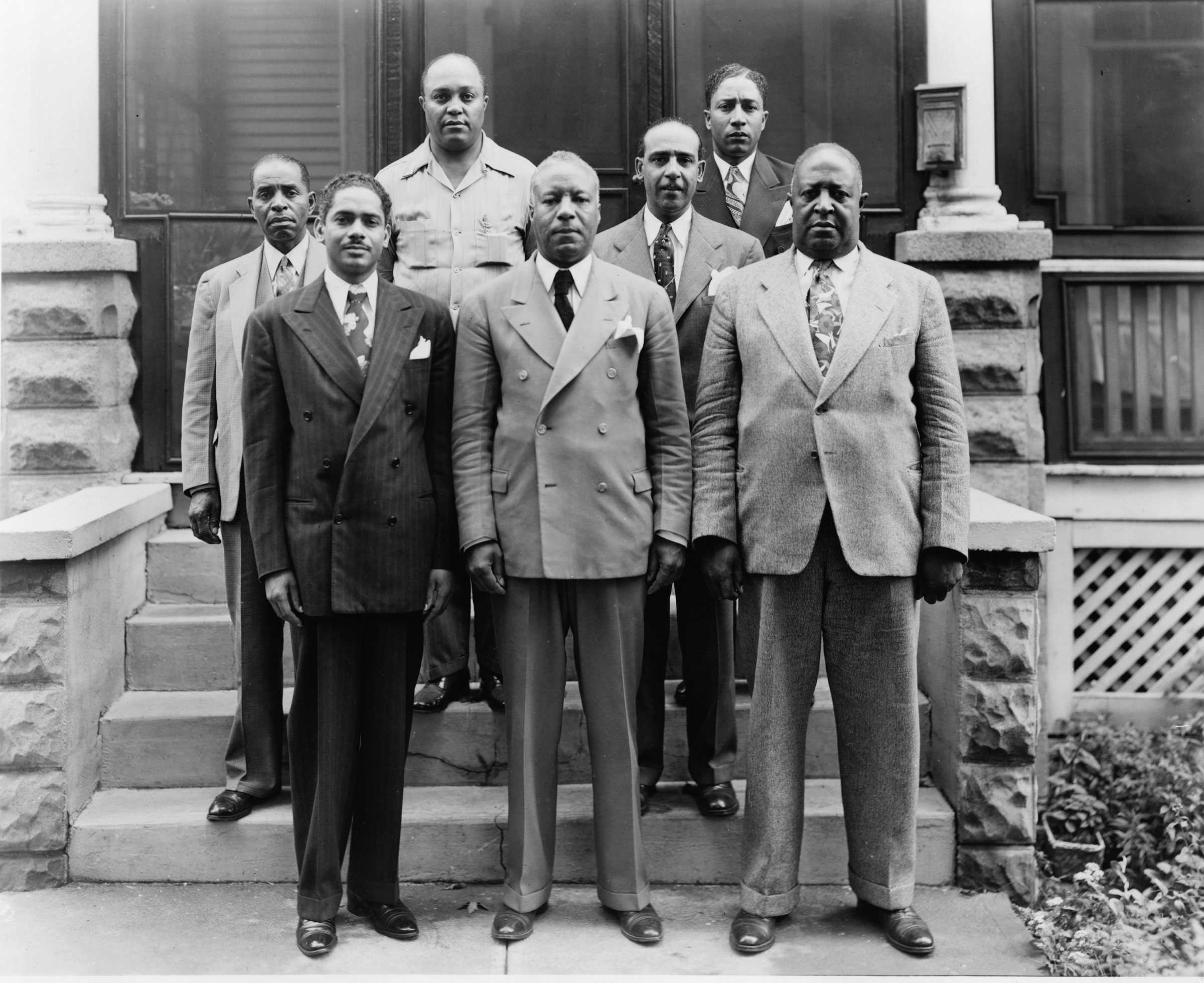 A black and white photograph of A Phillip Randolph and Milton Webster with five other African American men. Dressed in suits, they stand on a stoop for the picture.