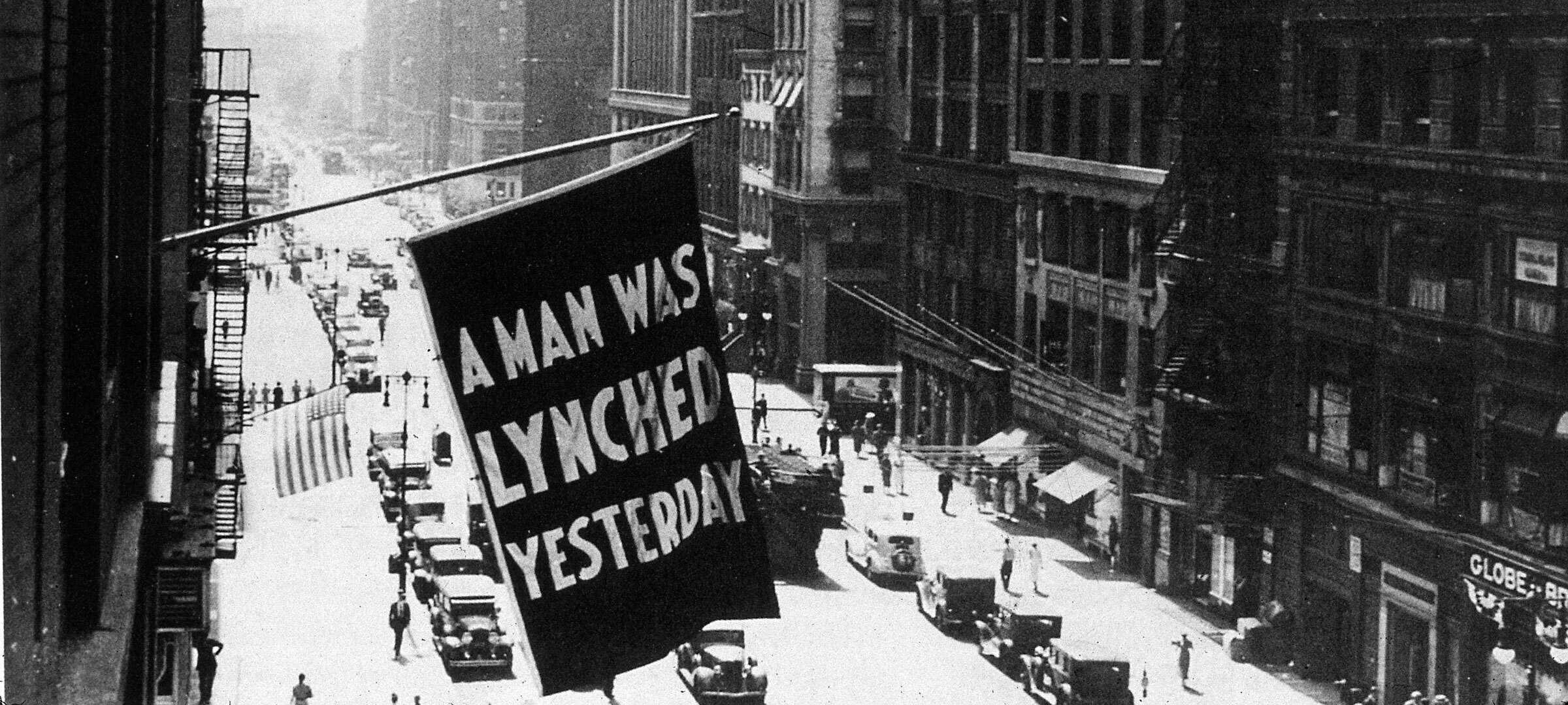 Black and white photograph of city buildings, one with a flag that says"A man was lynched yesterday".