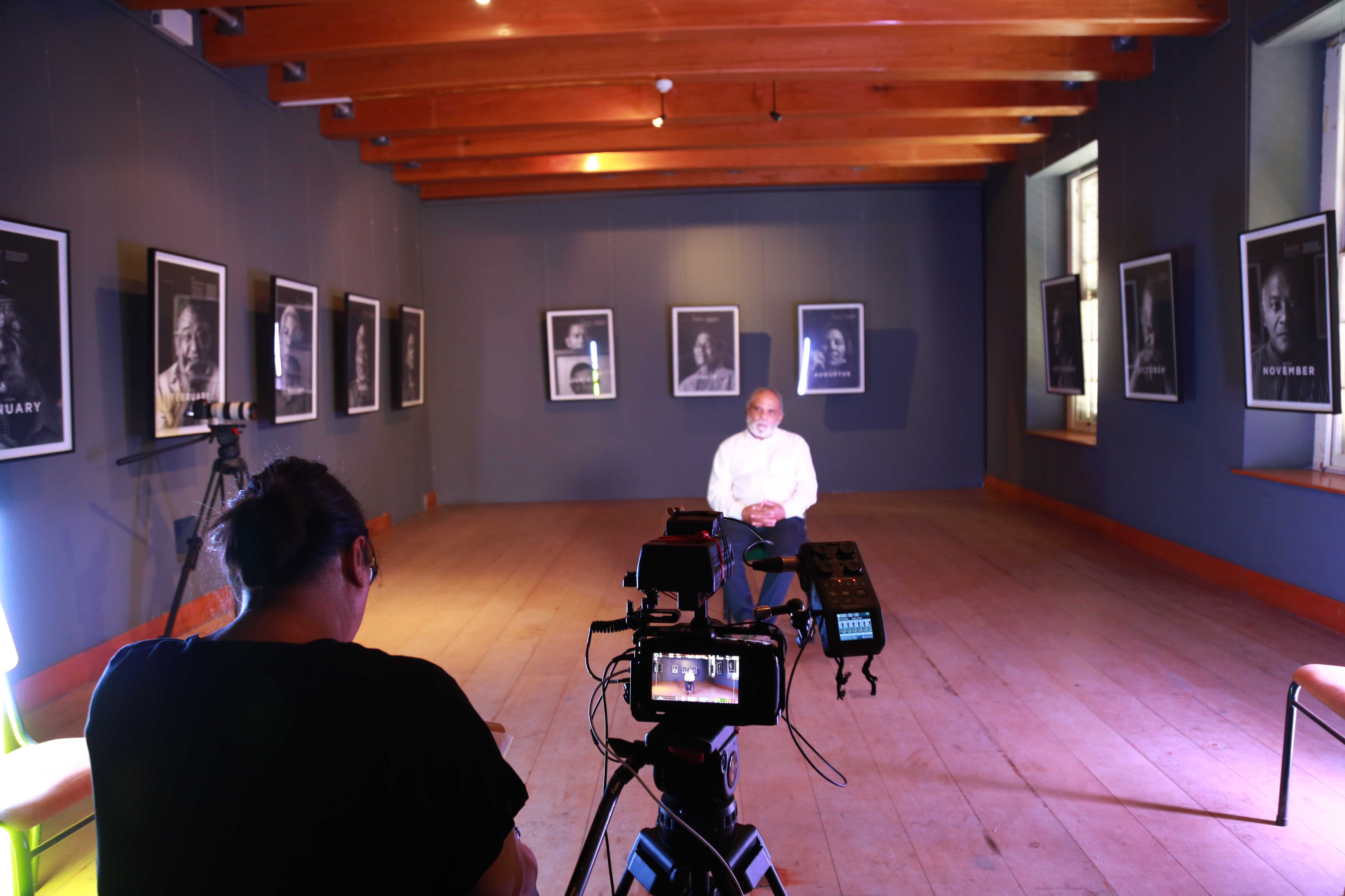 Roderick Sauls sits for an interview in a larger room. A camera is focused on him with director besides it.