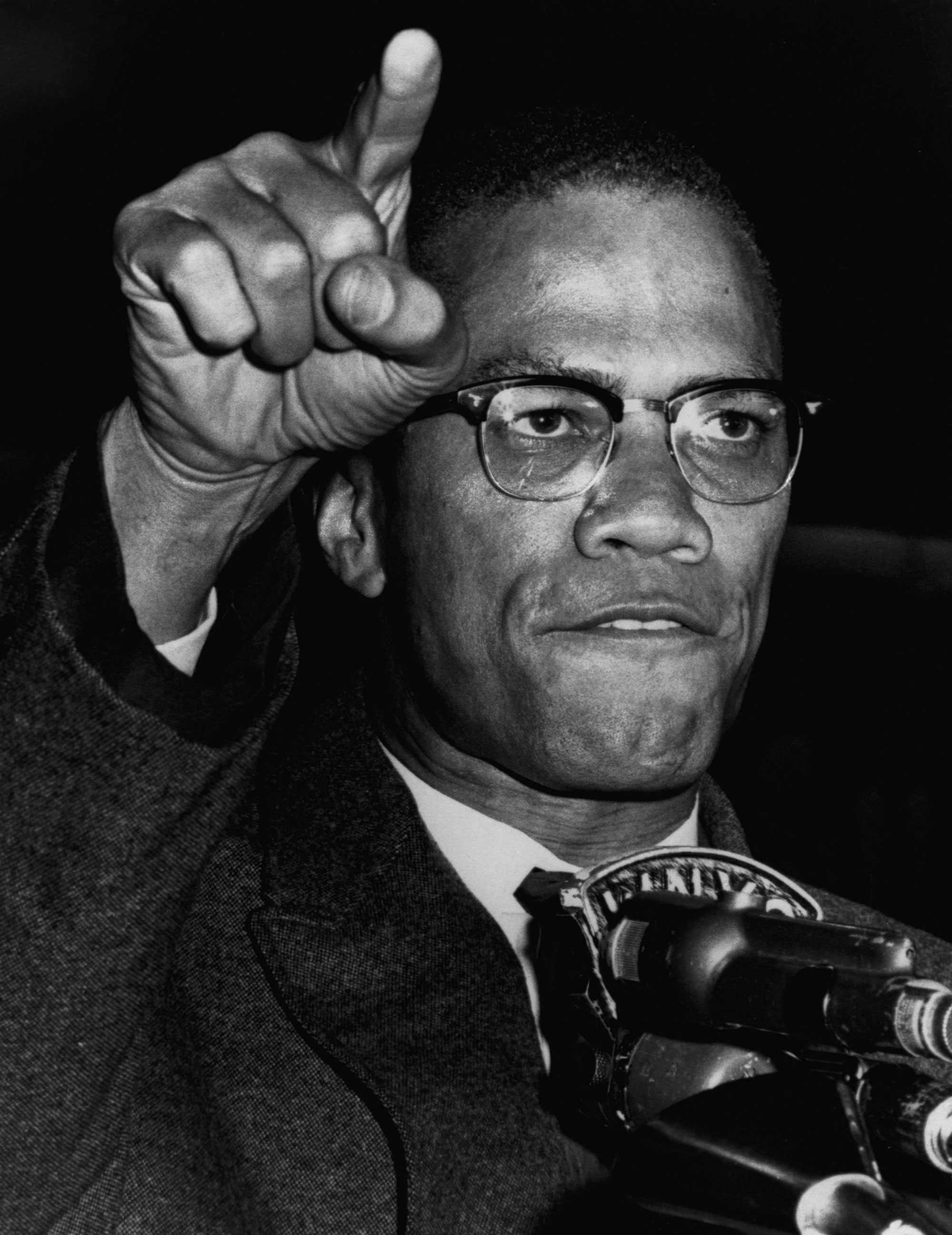 A black and white close up photo of Malcolm X speaking. He is dramatically pointing into the crowd, as he lightly bites his lower lip as he speaks.