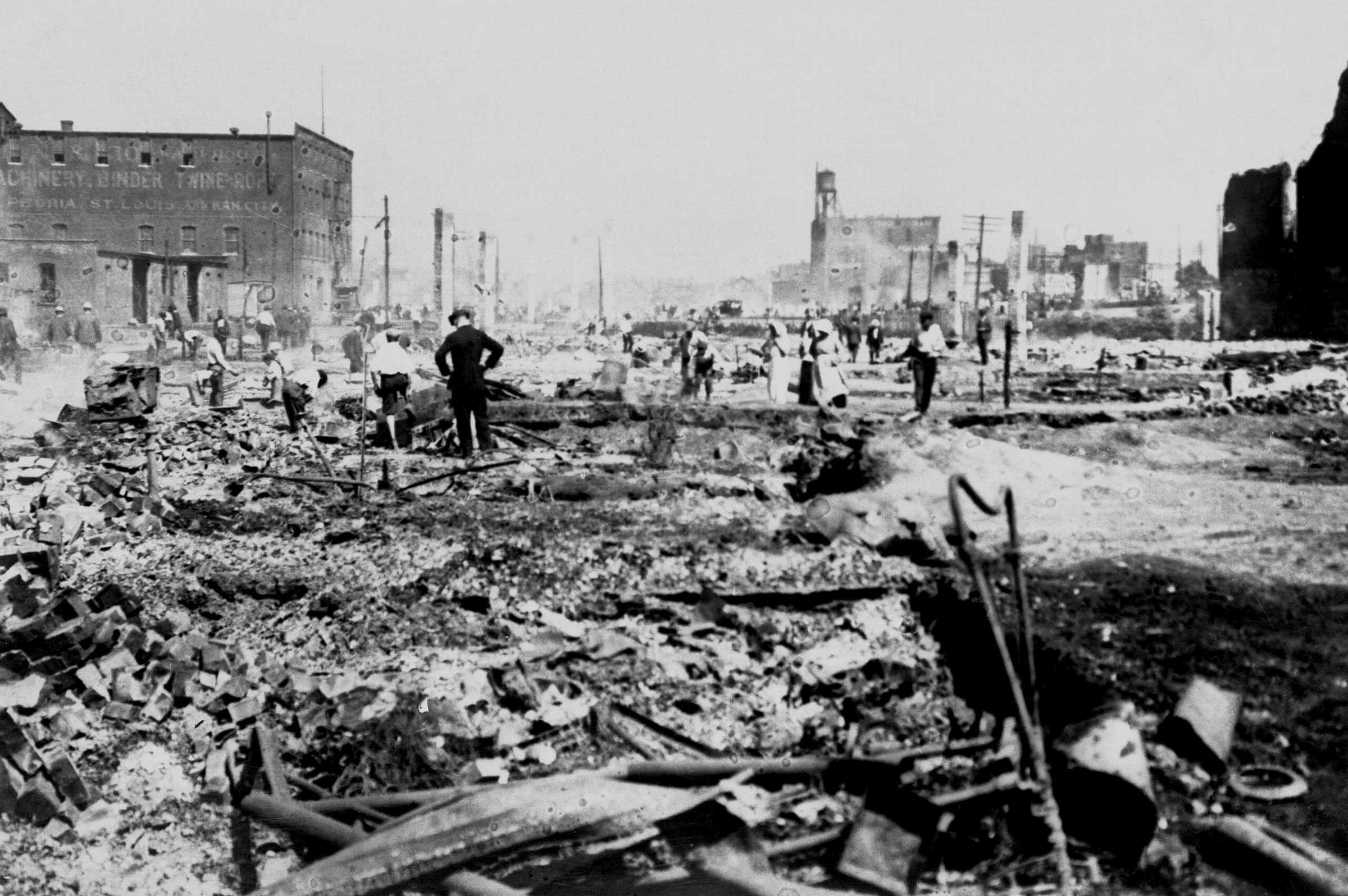 Black and white photograph of a destroyed city block with people in the distance going through rubble.