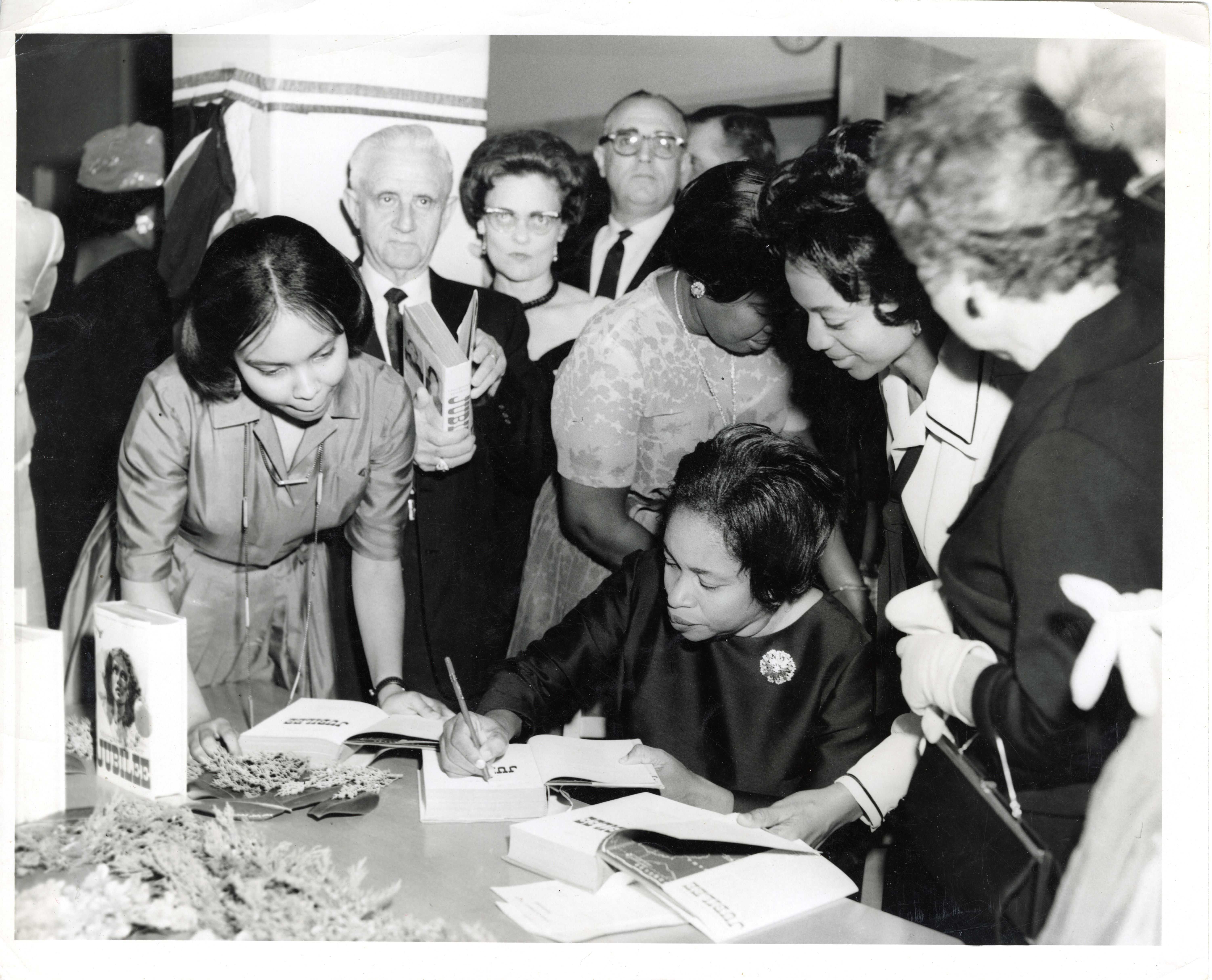 Black and white photo of a book signing event, with a seated individual (Margaret Walker) signing books at a table surrounded by people holding or observing copies of the same book.