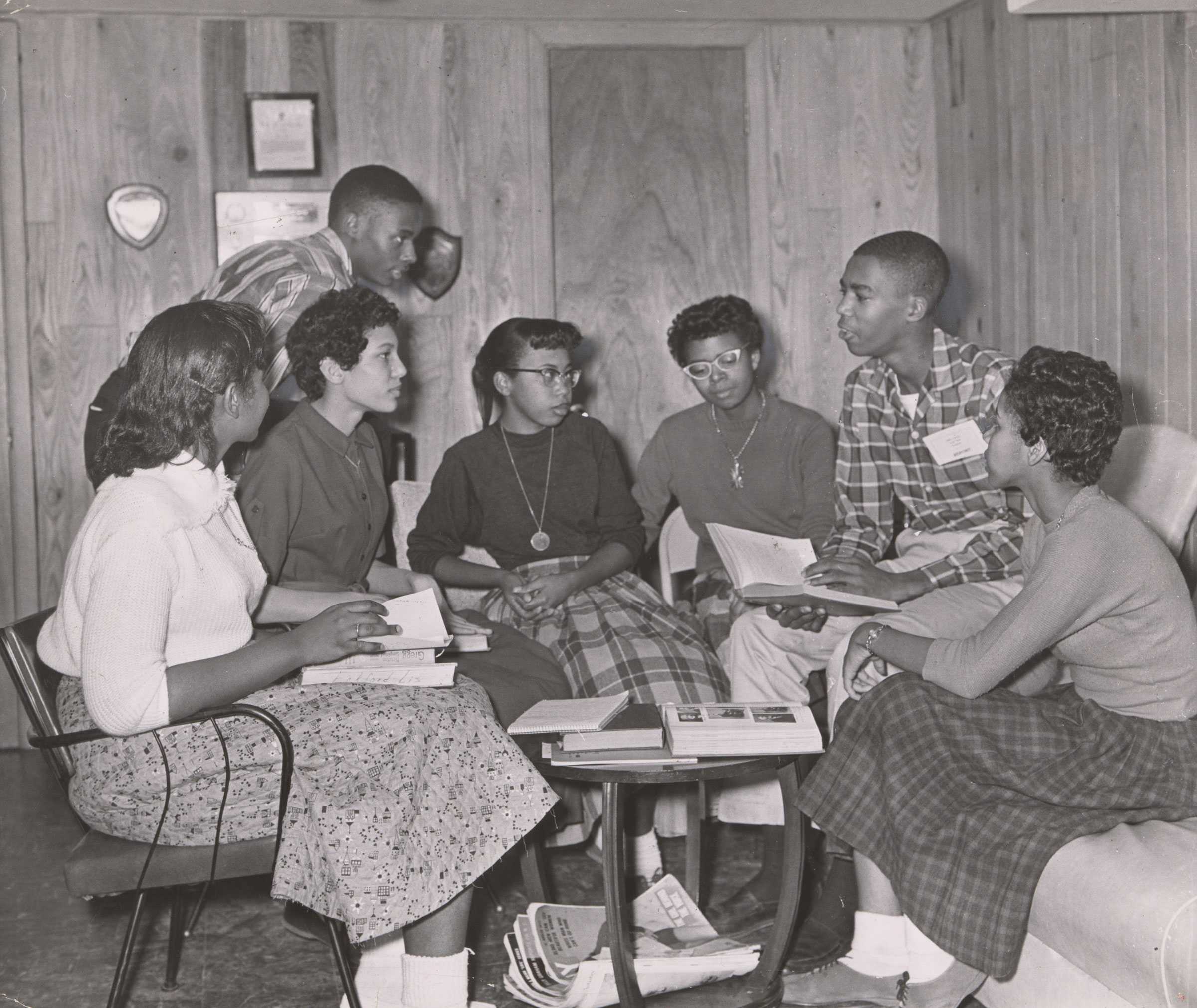 A black-and-white photograph of seven of the Little Rock Nine—Melba Patillo, Carlotta Walls, Jefferson Thomas, Elizabeth Eckford, Thelma Mothershed, Terrence Roberts, and Gloria Ray—studying around a table at Daisy Bates' home.