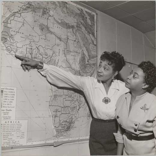 Black and white photograph of Etta Moten Barnett and another woman examining a large map of Africa hanging on the wall. Wearing a long sleeved white blouse with a brooch at the neckline and a grey straight skirt, Etta Moten Barnett stands to the side of the map and points to the country of Liberia with her right arm outstretched. The woman standing next to her wears a light grey dress with double breasted black button up the front and a black belt.