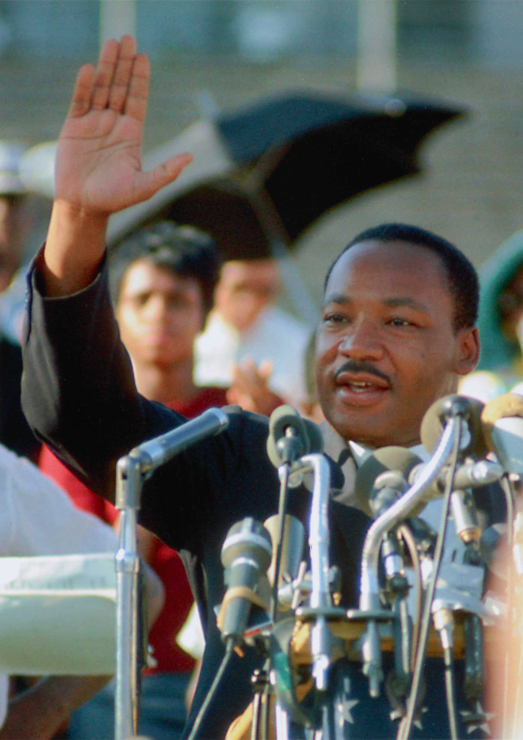 A color photograph of Dr. Martin Luther King, Jr. at a podium with multiple microphones. He is holding his right hand in the air, palm outward.