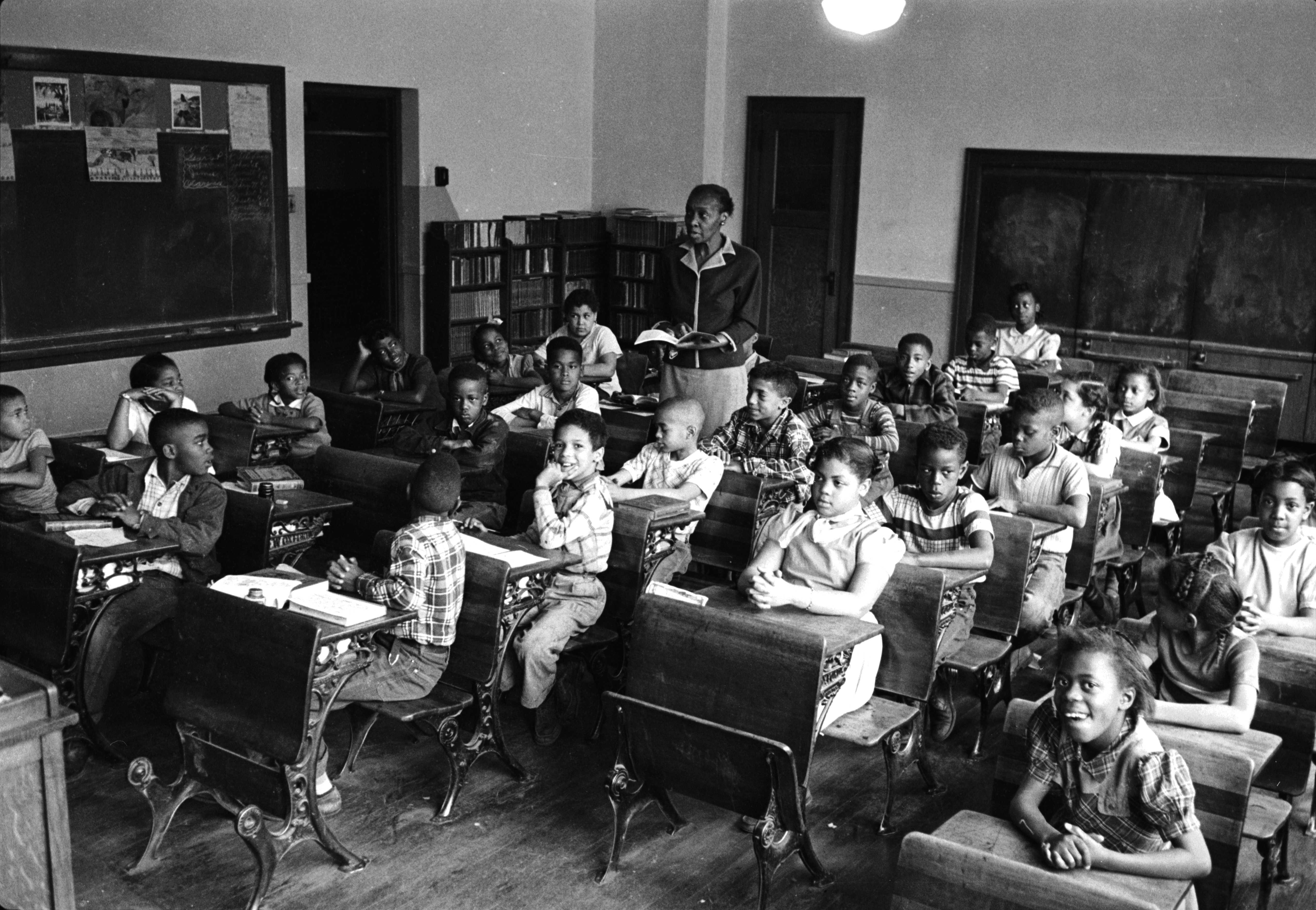 Black and white photograph of classroom with children seated at desks.  The teacher reads from a book while standing in the middle of all the desks.