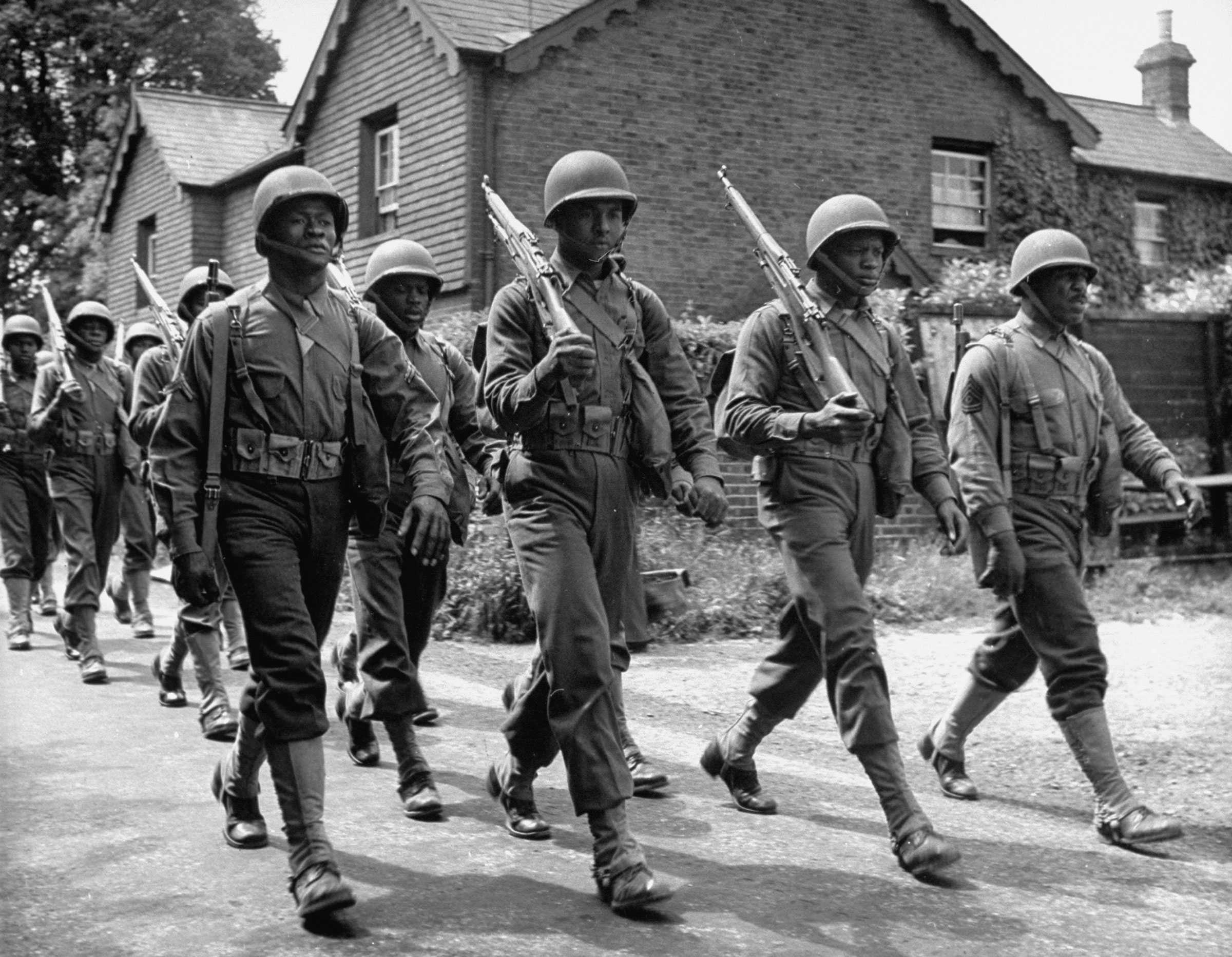 Black and White photograph of African American soldiers marching in formation in a small village.