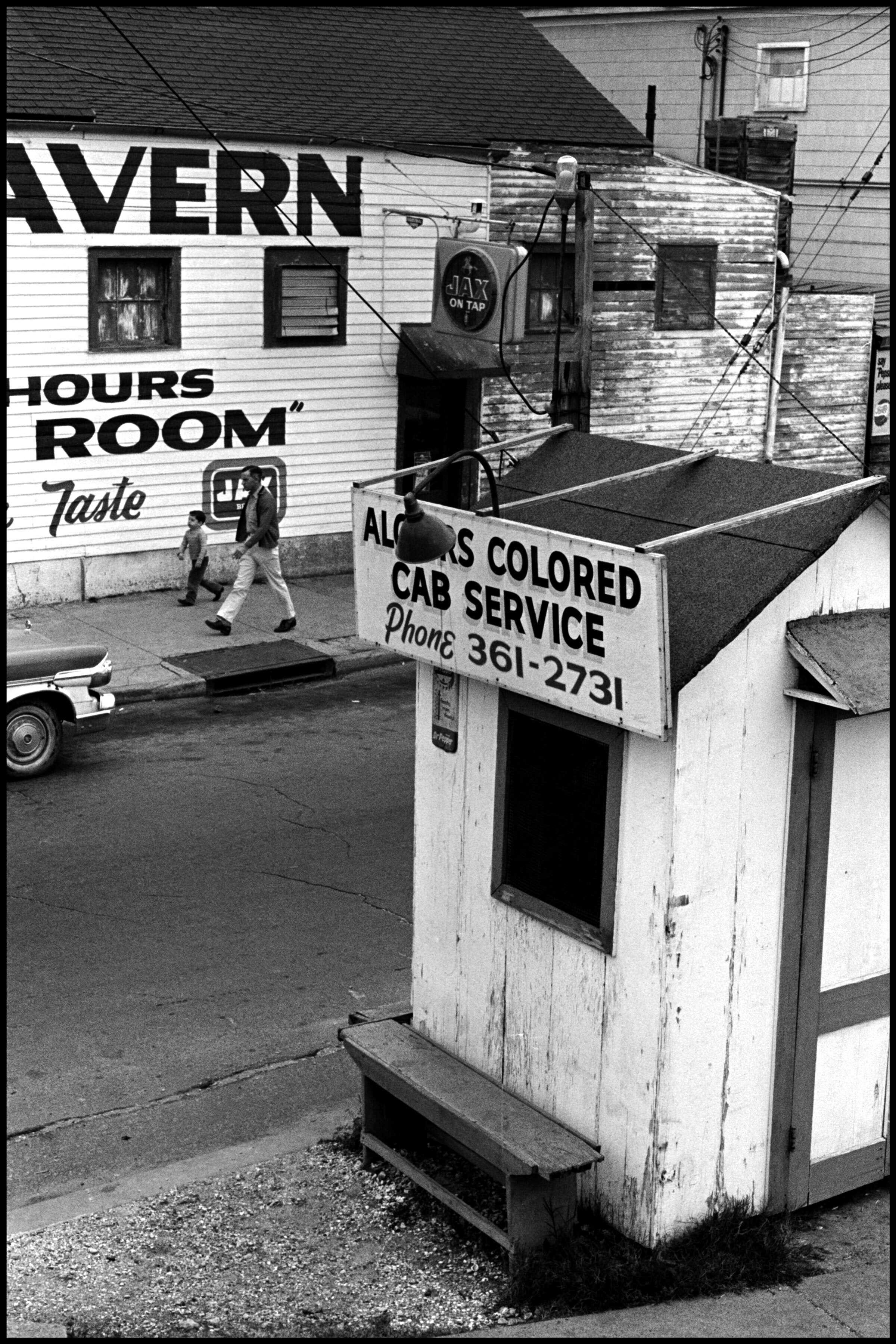 A black and white photograph of a cab sign on a street. Someone is walking on the sidewalks behind the sign.