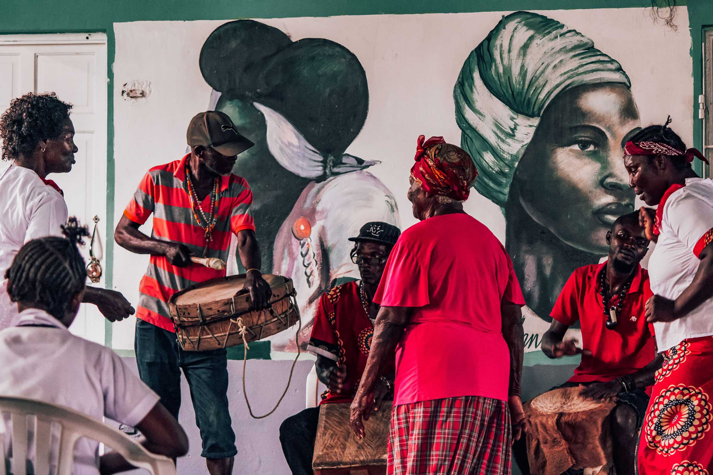 3 drummers play infront of group of women. A mural of two marooned women.