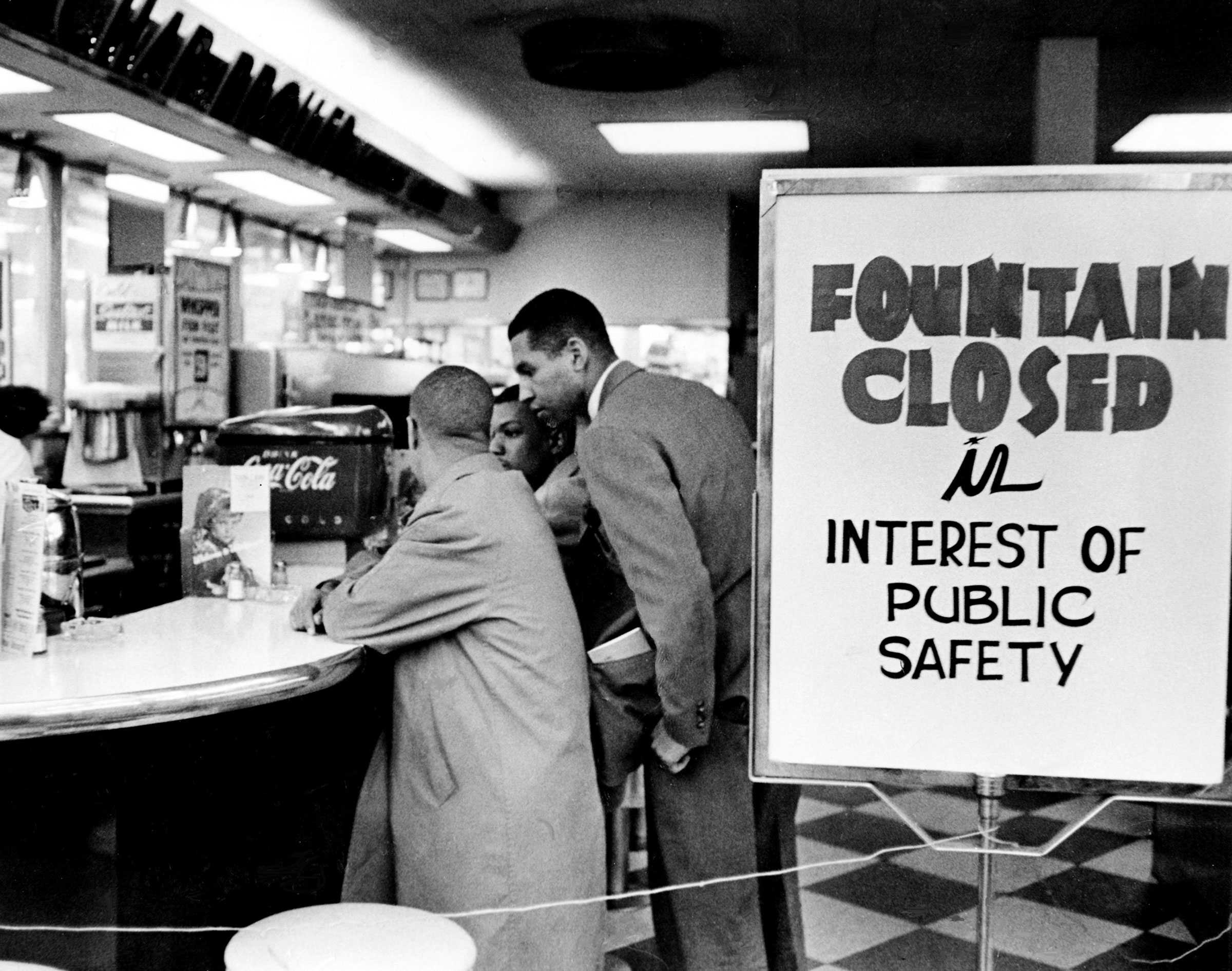 A black and white photograph of 2 people sitting at a Walgreens. They sit behind a sign that says "Fountain Closed in interest of public safety"