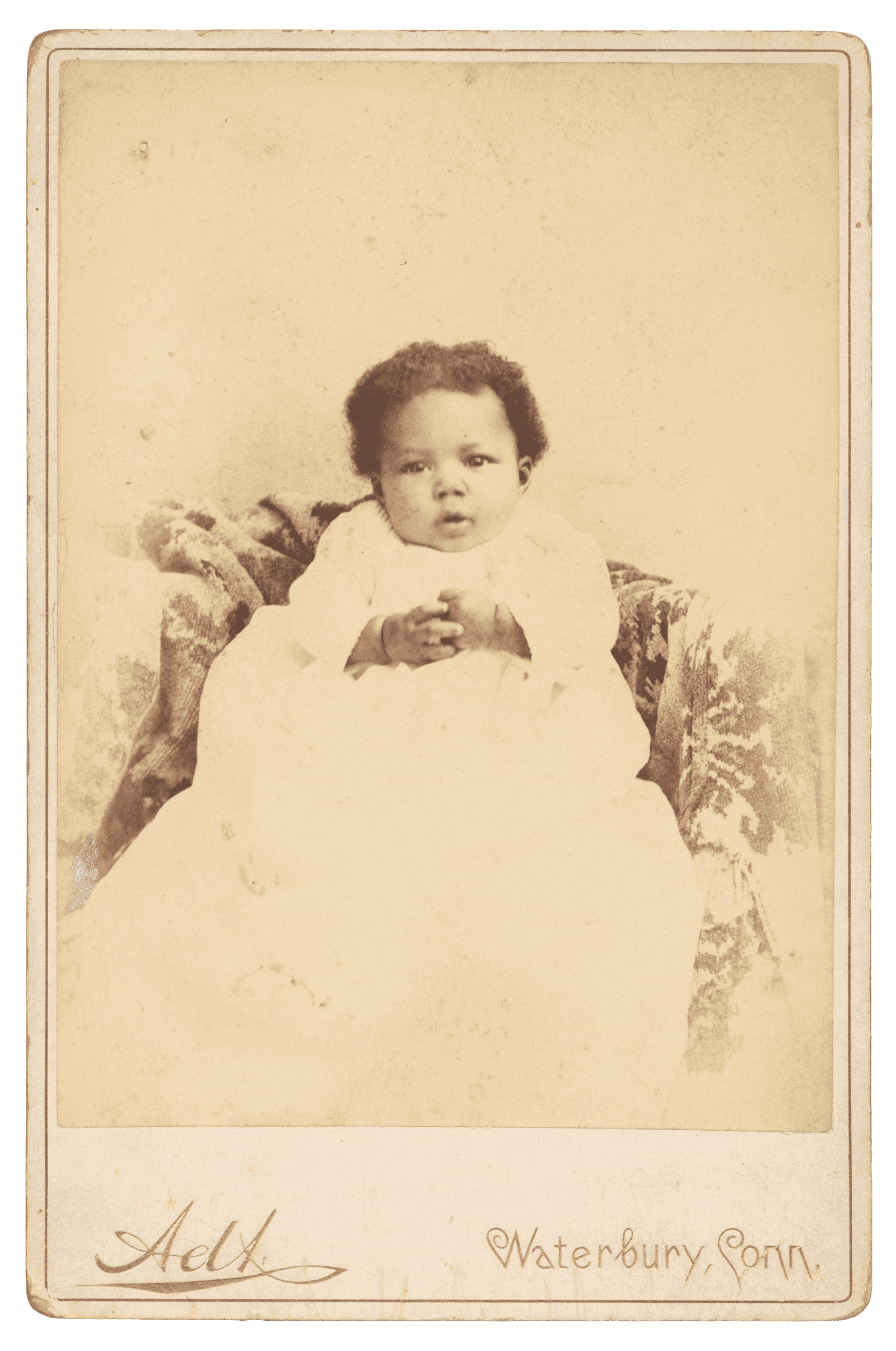 A cabinet card with a photo of an infant in a long christening gown seated on a patterned blanket.