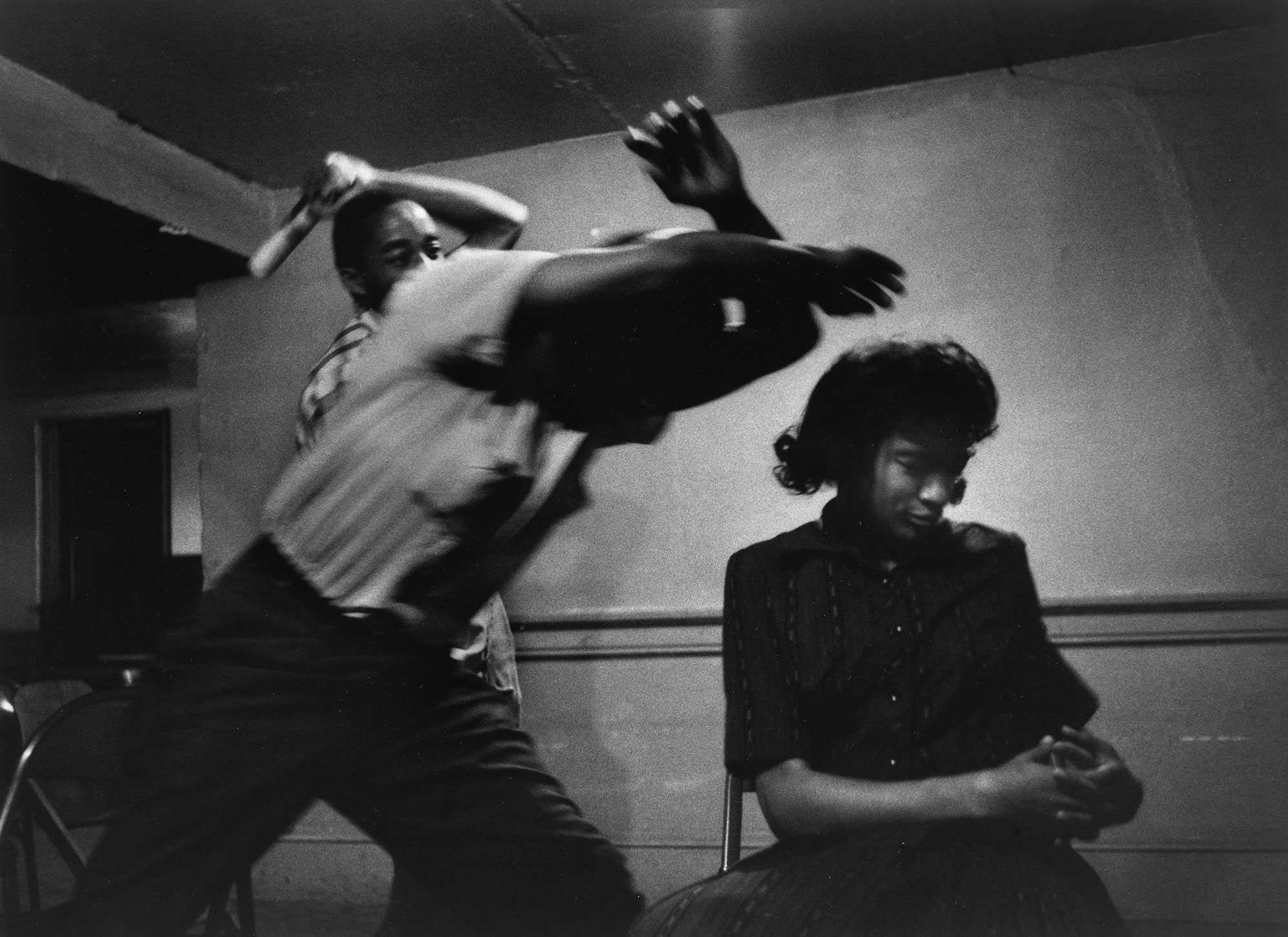 A black and white photograph of woman sitting down as two men hit her from behind as part of Passive Resistance Training.