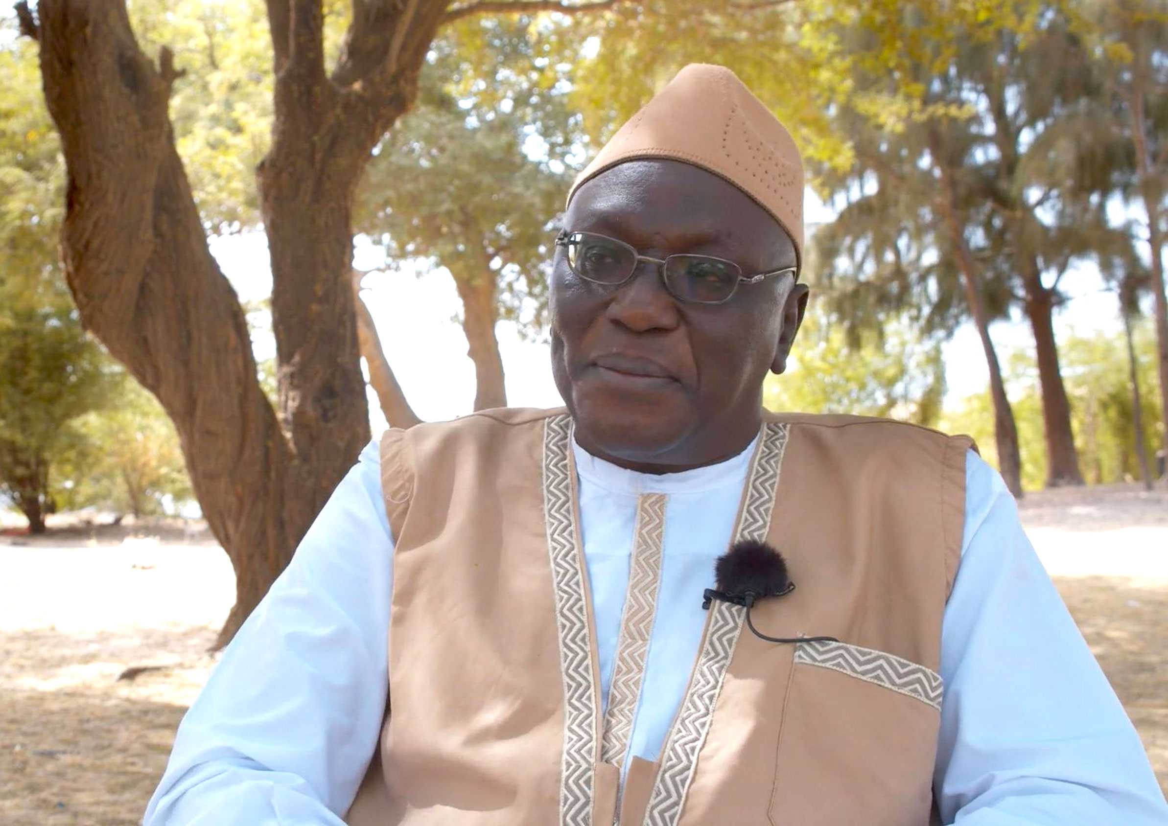 Mamadou Ba sitting for an interview while wearing a cream dashiki suit while wearing a clip on microphone.