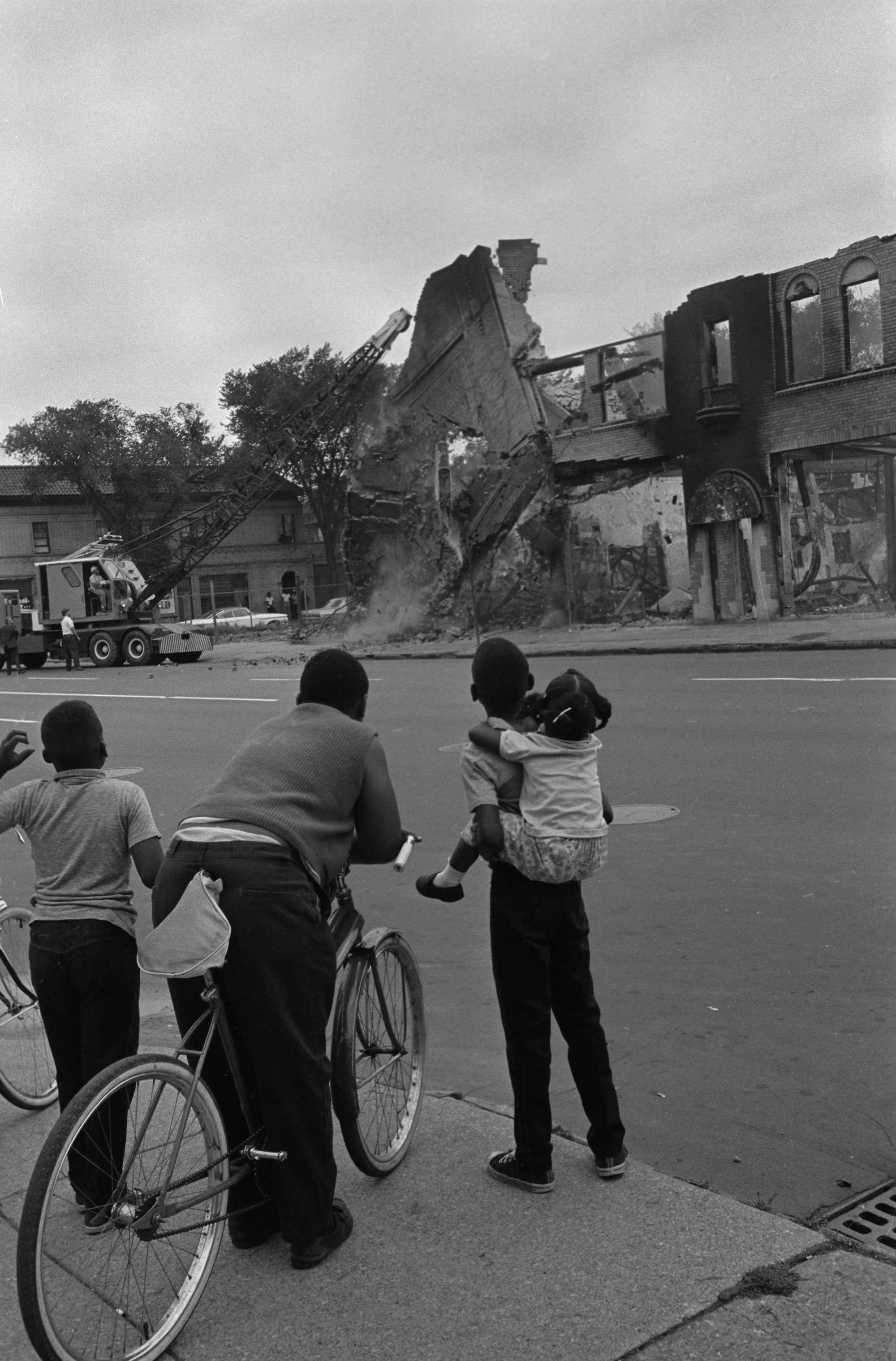 A black and white photo of a group of Black children watch across the street  as crane knock down a burned building.