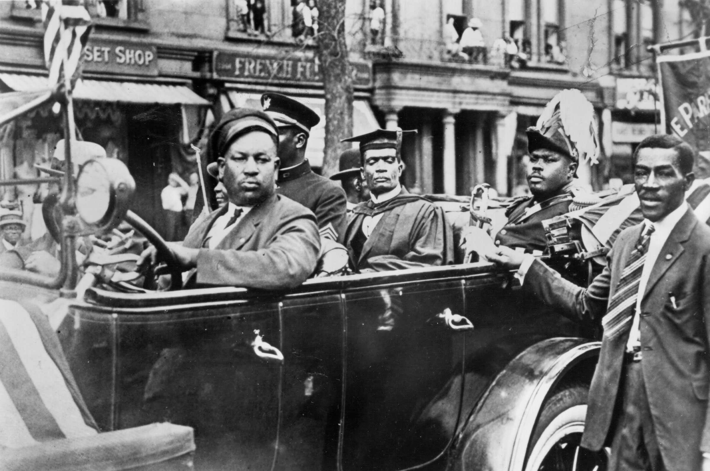 Black and white photograph of men seated in an open car.  They appear to be participating in a parade.  One man is in academic regalia, another is military clothing and a third in military clothing from a non-US division.