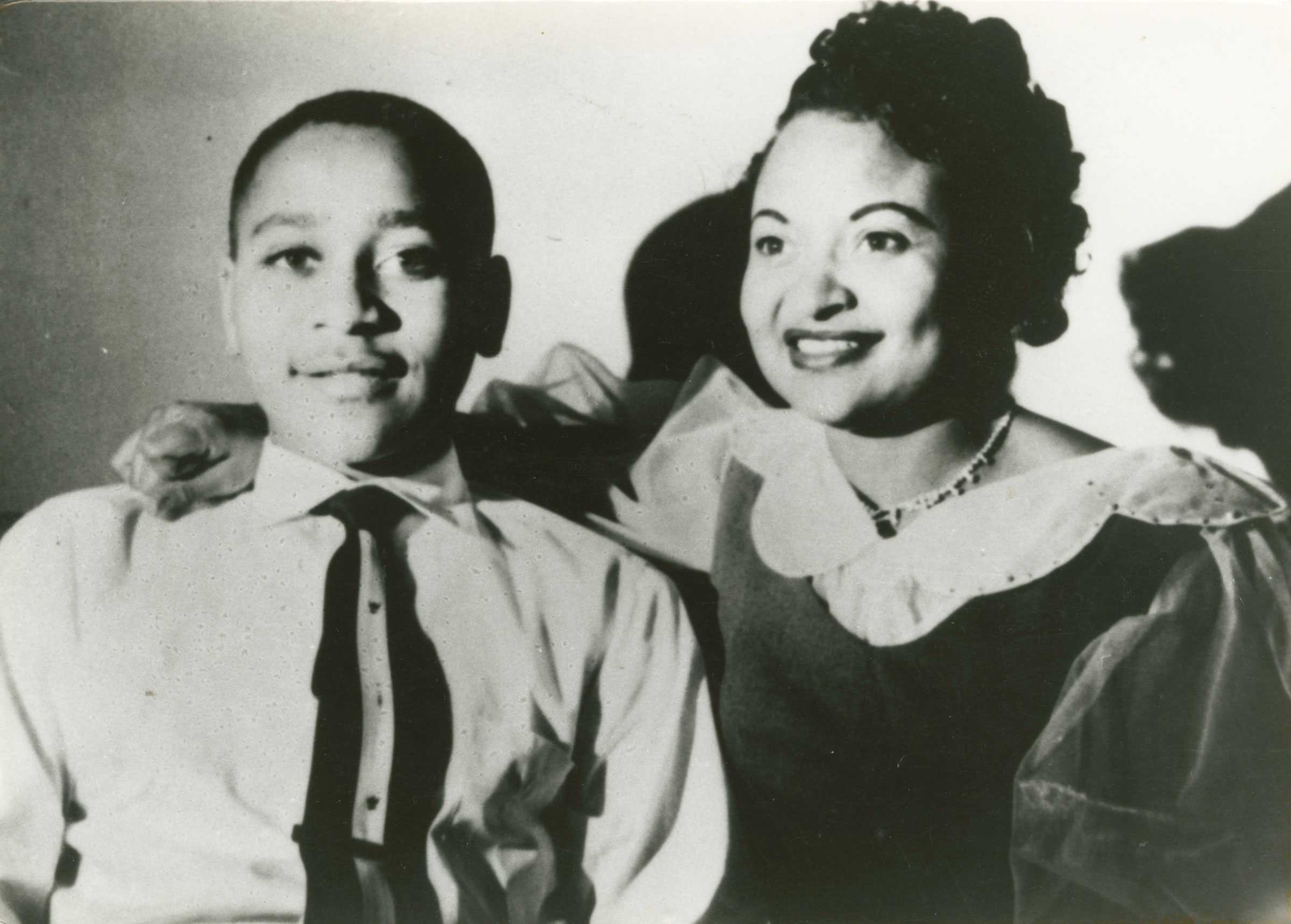 Black and white photo of Emmett Till with his mother, Mamie Till Mobley. He wears a white shirt and tie; she sits beside him in a dark dress with a ruffled collar.