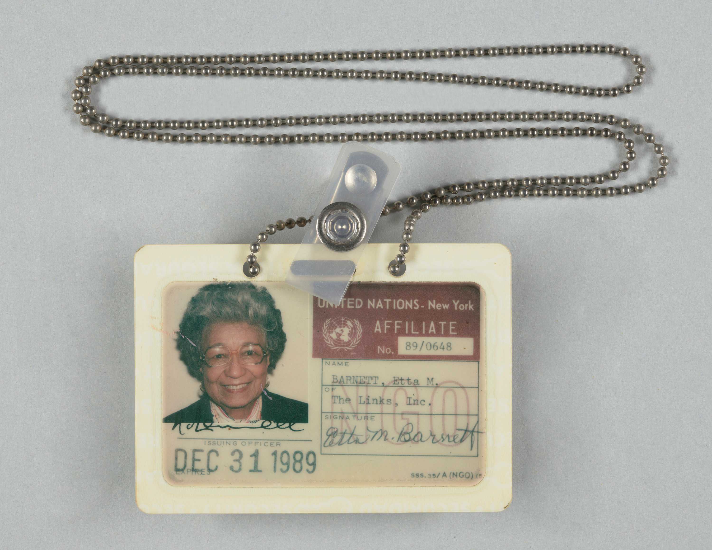 United Nations ID card belonging to Etta Moten Barnett. It is a plastic laminated card with a color photograph of Etta Moten Barnett in the top left hand corner. In the photograph Etta Moten Barnett is depicted from the shoulders up. She is wearing a black top, a pink scarf around her neck and glasses.