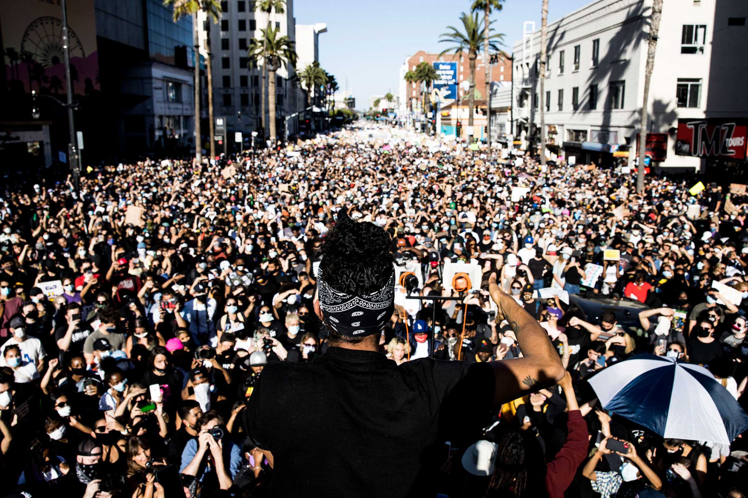 Janaya "Future" Khan, stands at the center facing away, their back to the viewer, addressing a large crowd at a Black Lives Matter protest.