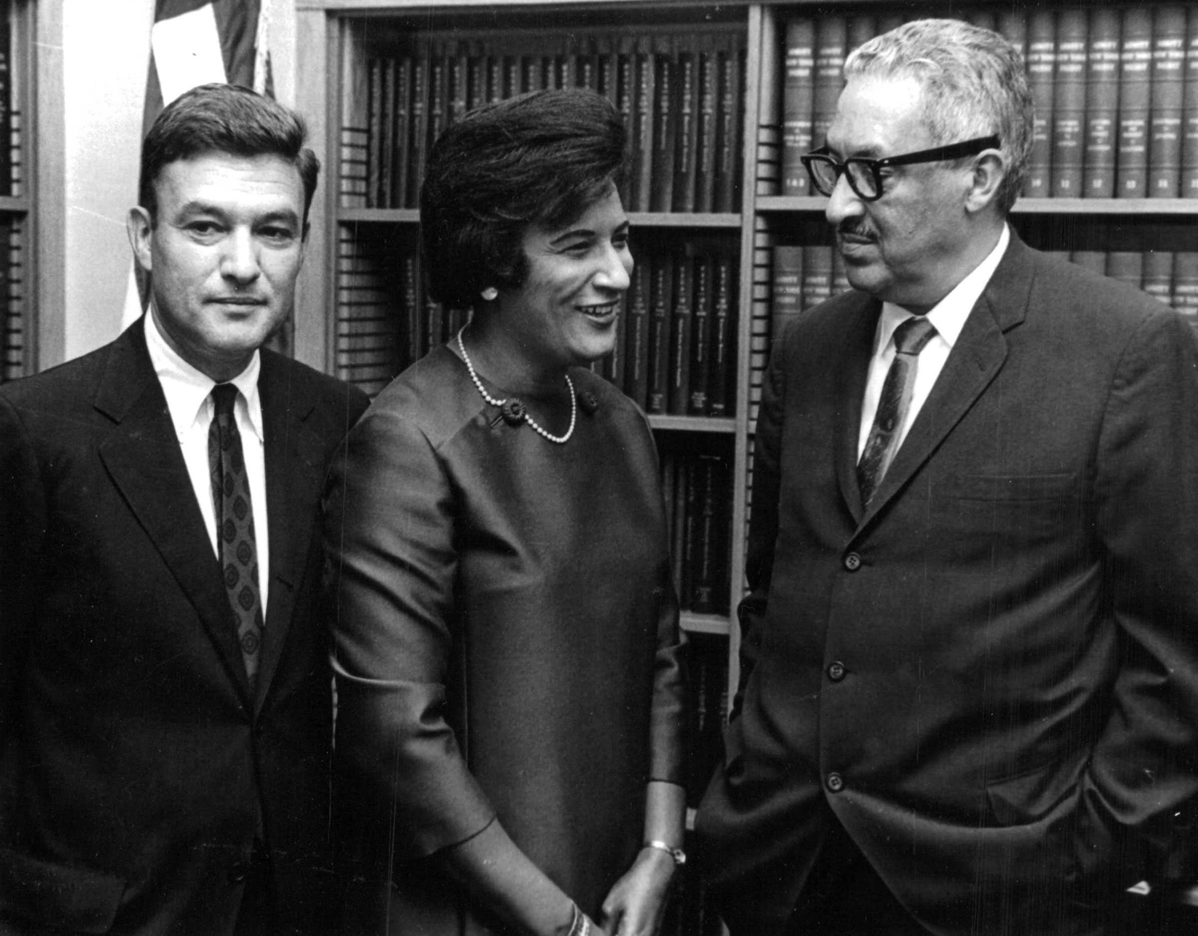 Constance Baker Motley, Thurgood Marshall and Jack Greenberg are gathered closely, talking to each other in front of a large bookshelf.