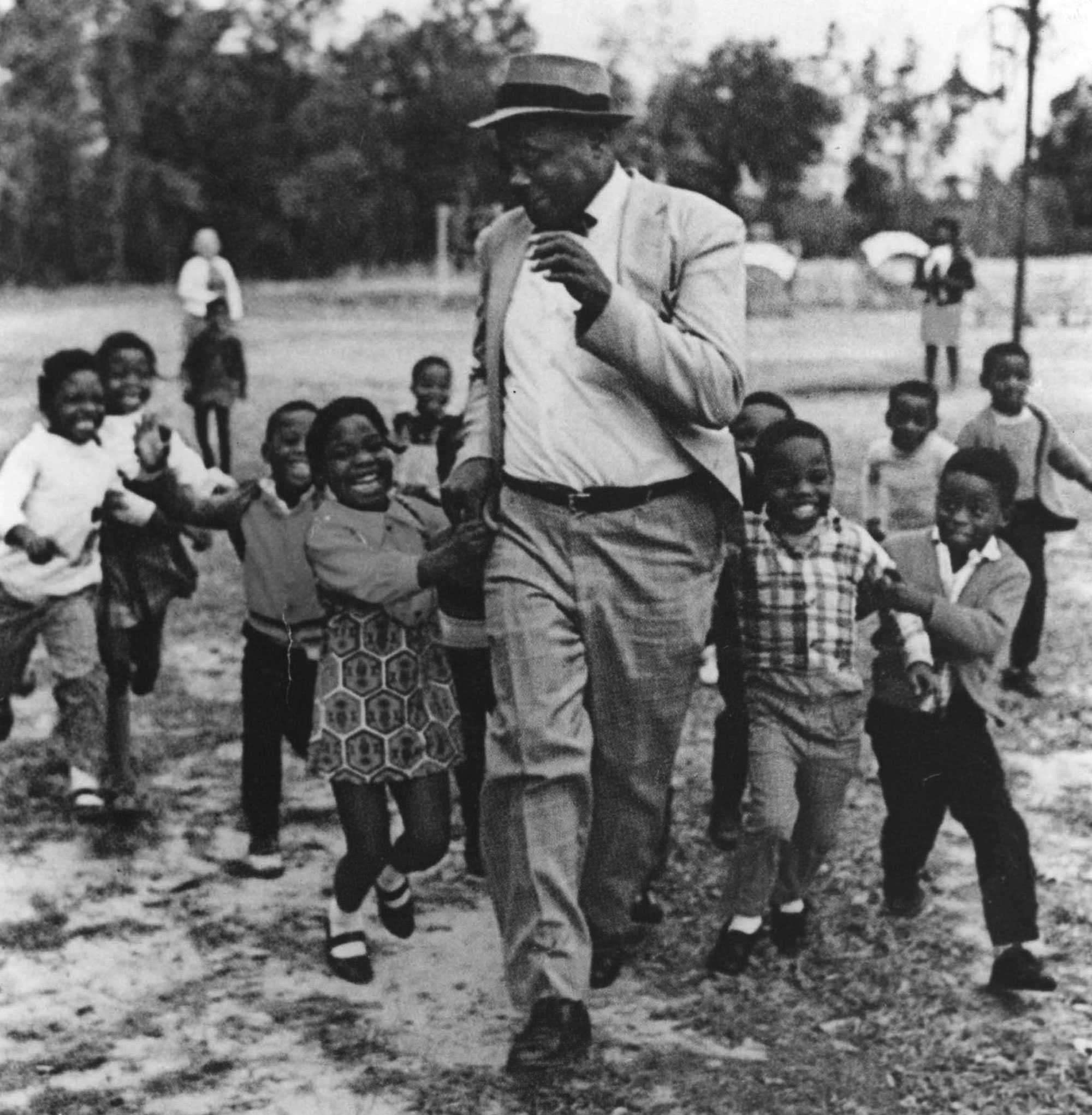 A black and white photograph of Esau Jenkins playing and running away from a group of young children. They laugh and smile as they chase him.