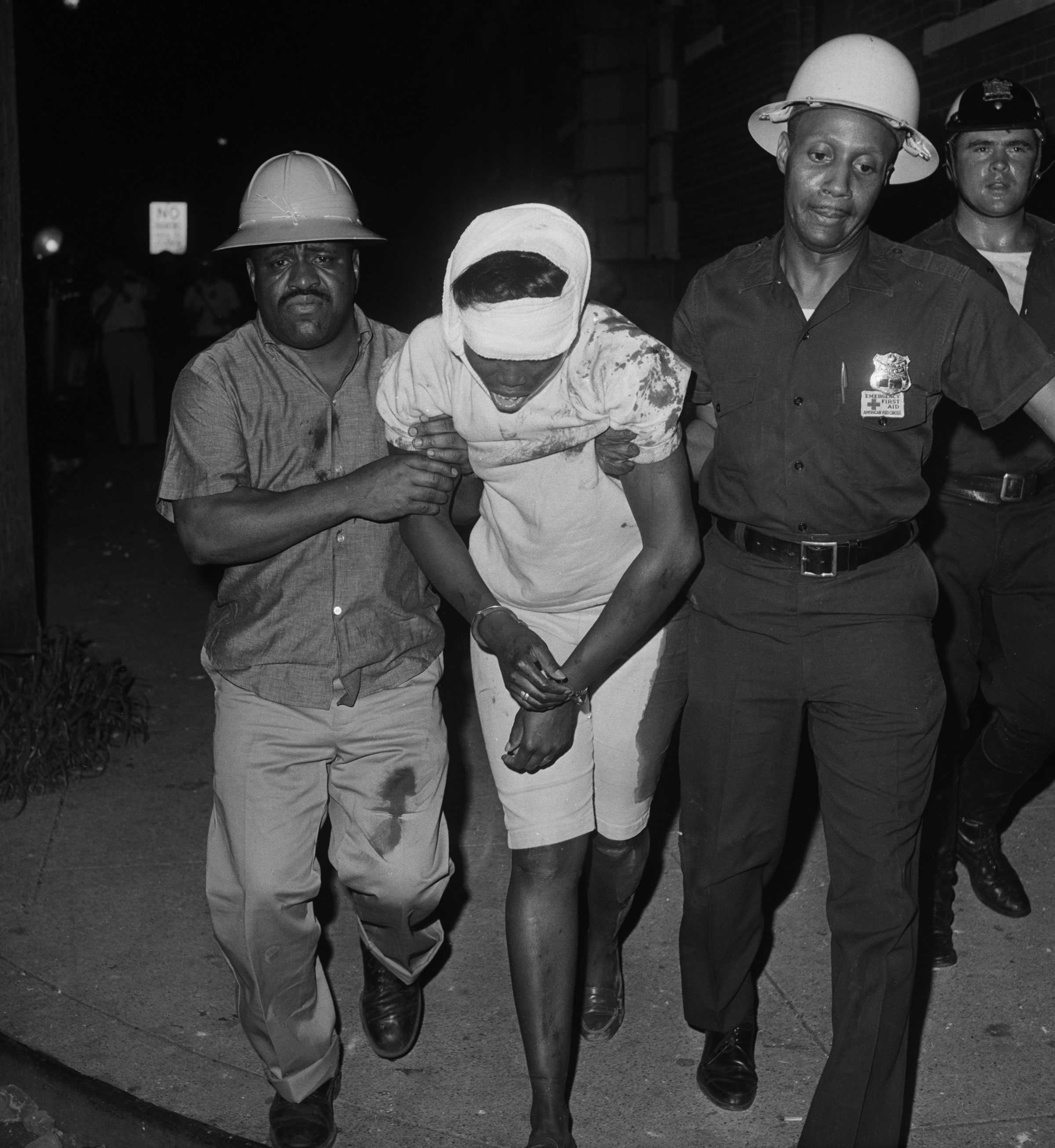 Two police officers remove a wounded woman. Her white clothes are stained with blood and her head is wrapped in white gauze. She is handcuffed.