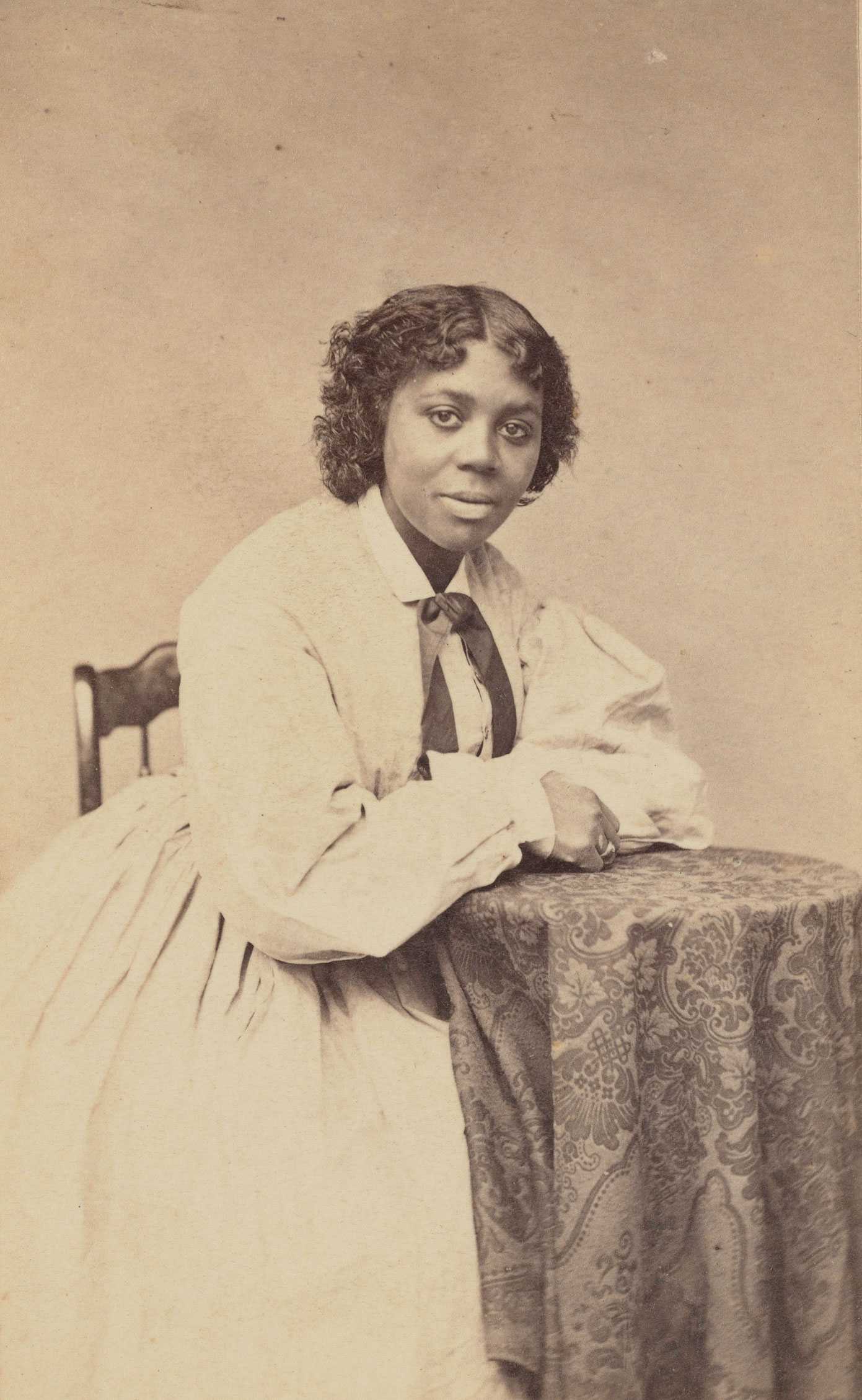 Sepia tone carte-de-visite of Edmonia Lewis seated in a chair, wearing a long dress with a ribbon at her neck.