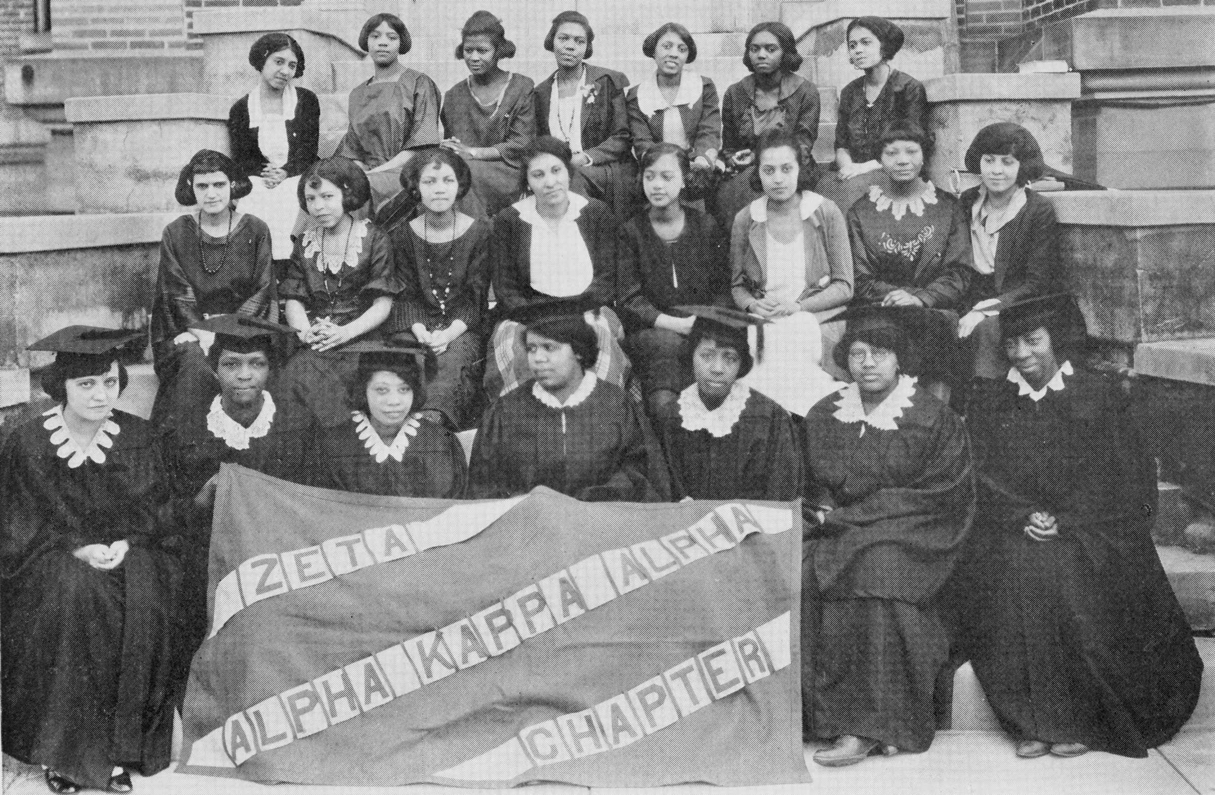 Black and white photograph of women seated on steps of a building, dressed in academic regalia (first row) holding a sign that says "ZETA / ALPHA KAPPA ALPHA / CHAPTER."  There are two more rows of women behind them dressed in fancy clothing.