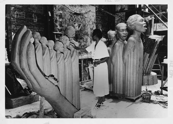 A black and white  photo of a woman working in a studio on a large sculptural piece.  The studio has brick walls and is cluttered with various tools and materials scattered around, indicating an active work environment. There are additional sculptures visible in the background.”