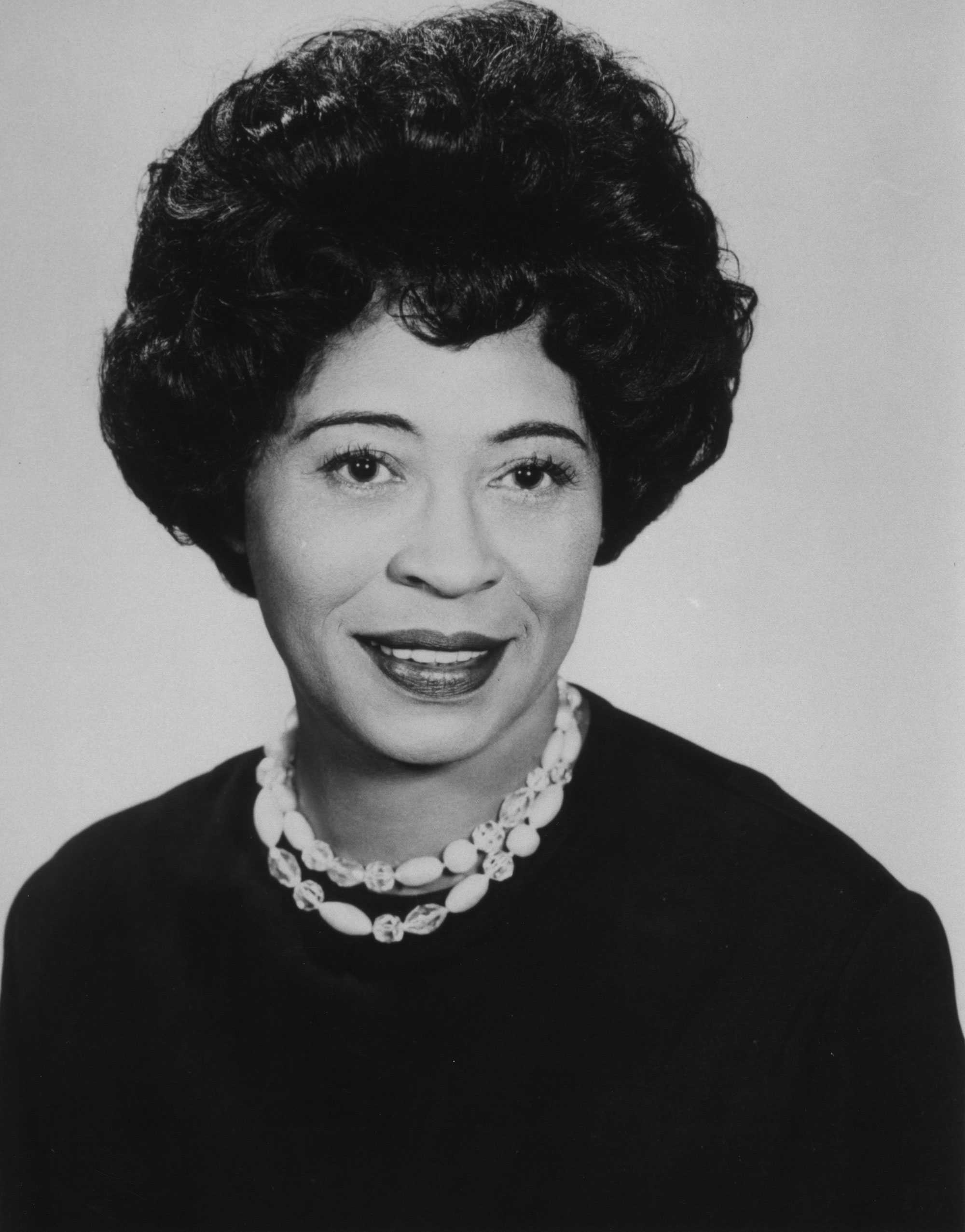 A black and white posed portrait of Daisy Bates. SHe is wearing a black shirt and chunky white beaded necklace. Her hair is short and curly.