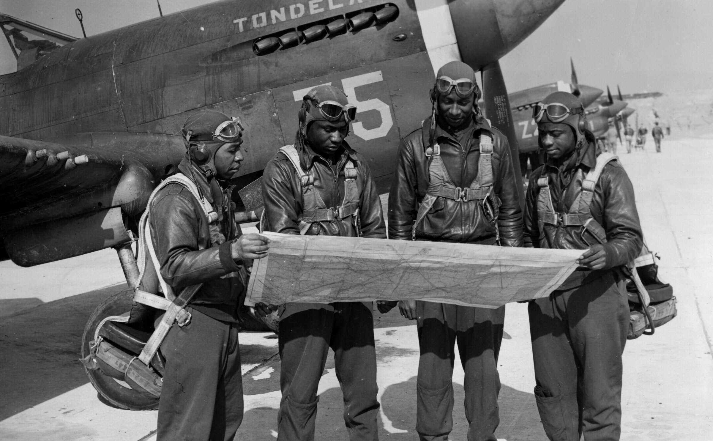 Black and white photograph of 4 airmen standing before a military aircraft studying maps.  They are all dressed in flight gear.