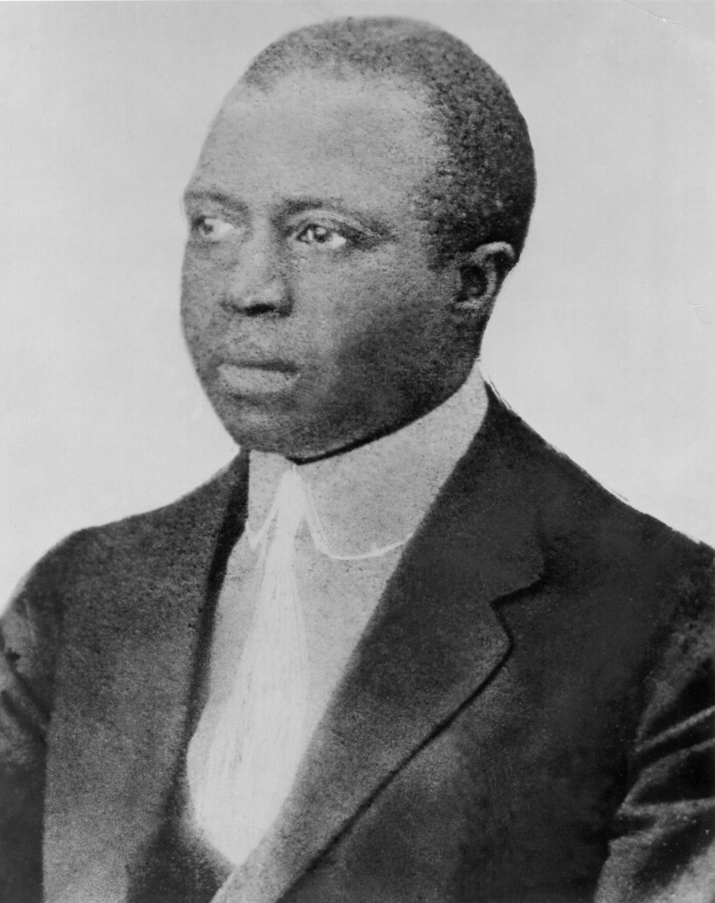 Shoulder length portrait of Black man dressed in a suit coat and white shirt and tie.