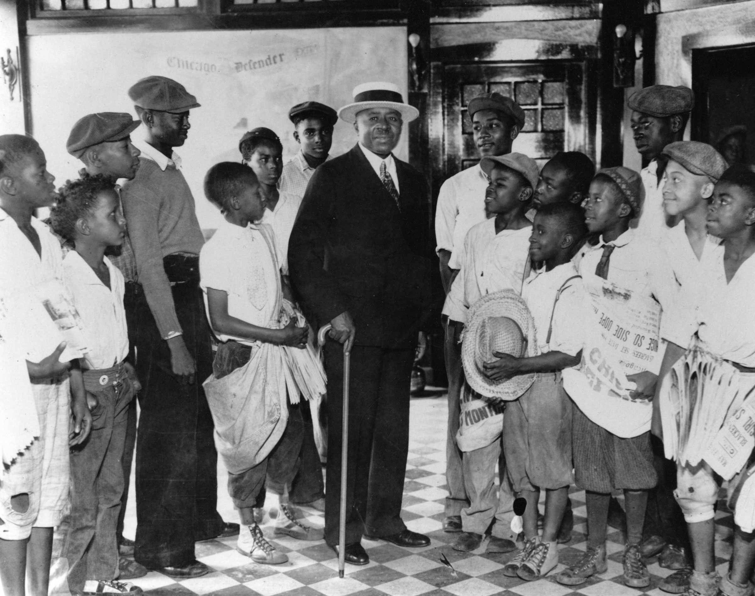 Black and white photograph of man dressed in dark suit holding a cane, surrounded on both sides by young men dressed in white shirts and caps  The young men appear to be holding newspapers in their hands.