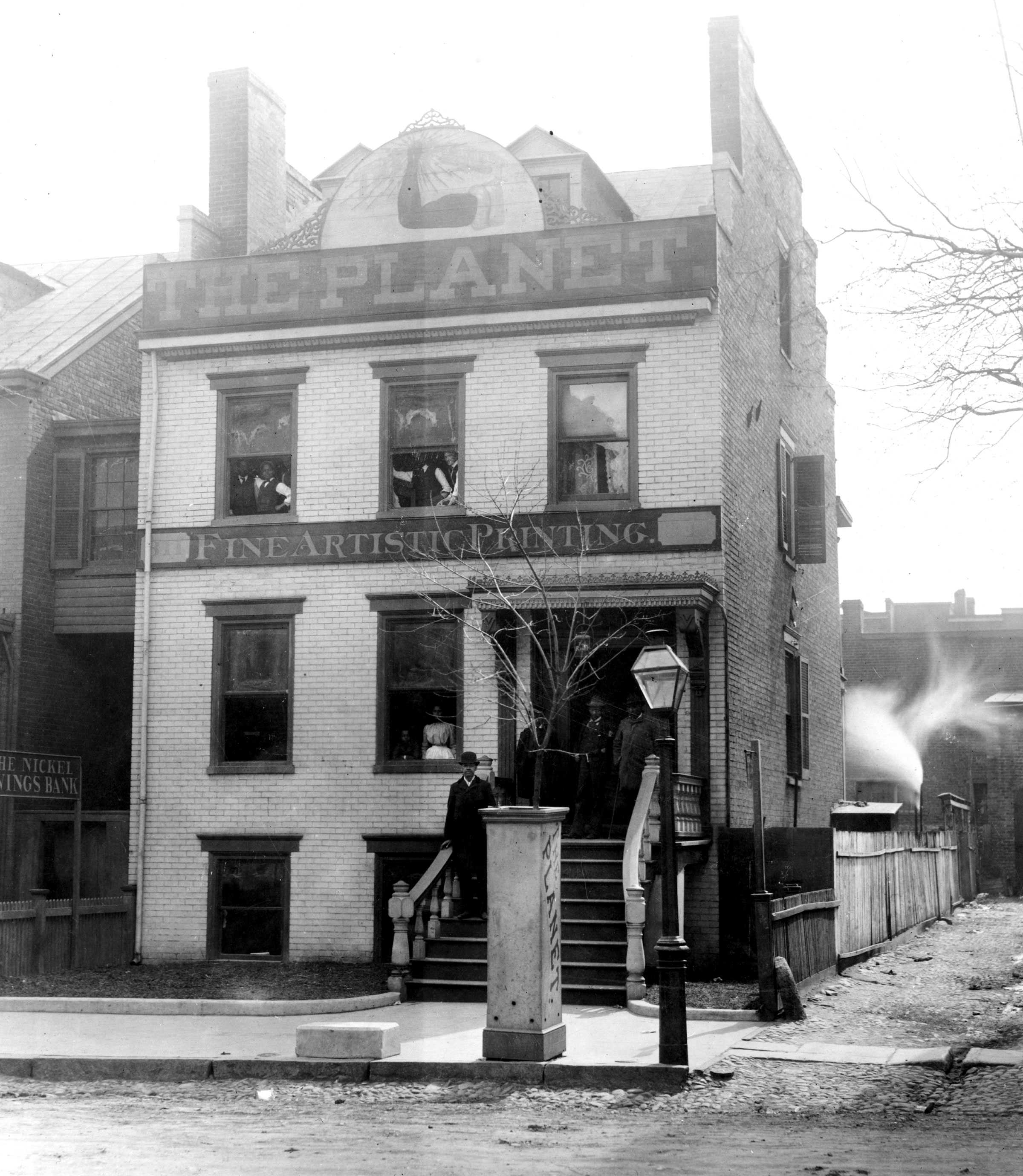 Black and white photograph of a 3 story white brick house.  The building is identified as "THE PLANET / FINE ARTISTIC PRINTING."   There are several figures visible standing on the front steps and in the windows.