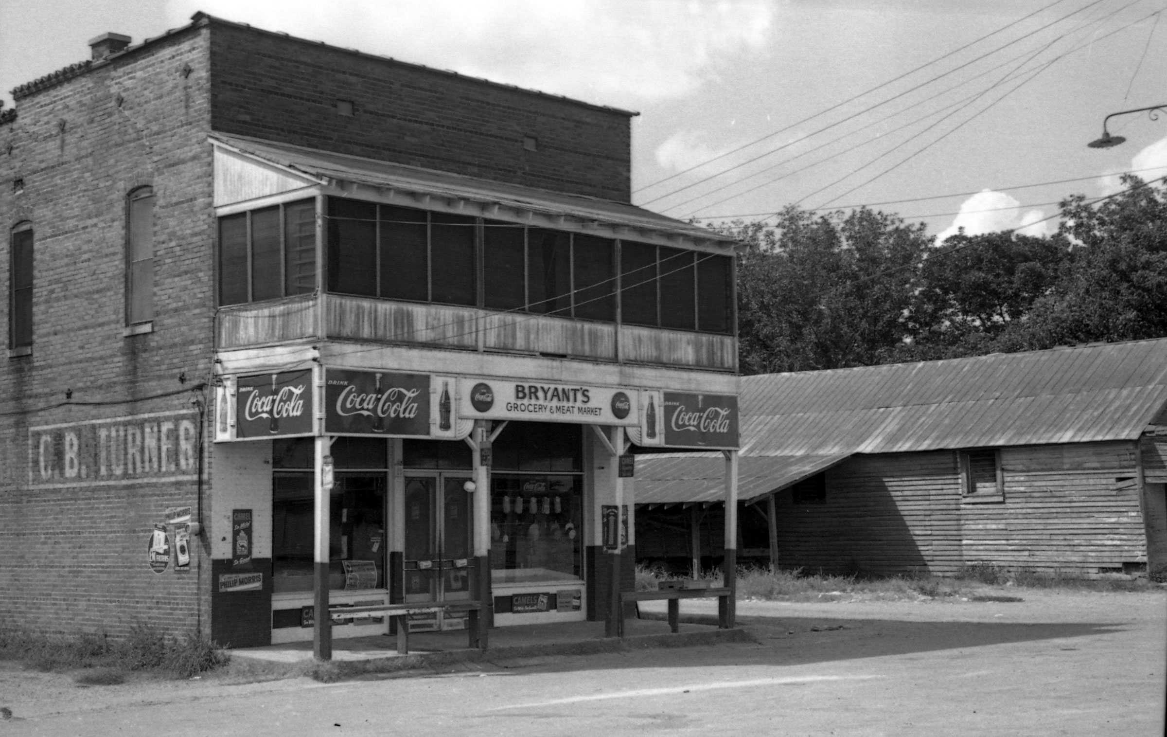 A black and white photograph of  the outside building of Bryant’s Grocery and Meat Market. It is a small brick building with a large store front.