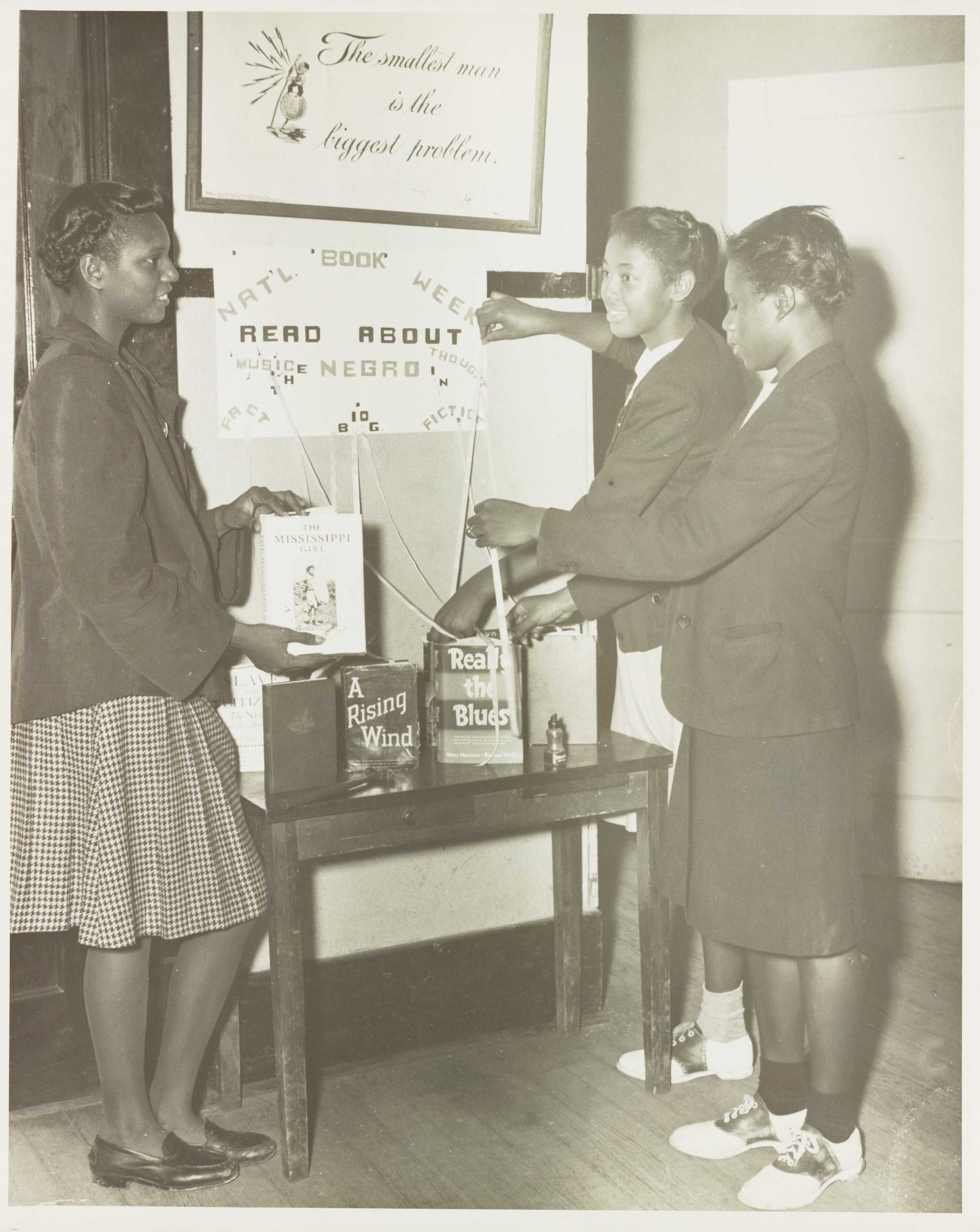 Black and white photograph of three young Black women standing before a small wooden table with books displayed on top.