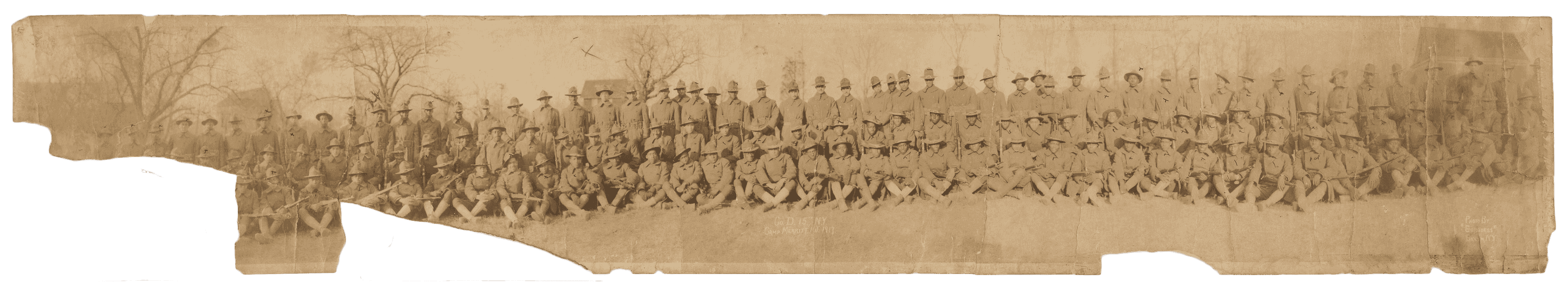 A worn and slightly torn, panoramic black-and-white photo of D Company, 369th Regiment, at Camp Merritt, NJ, in 1917. Soldiers in uniform with rifles.