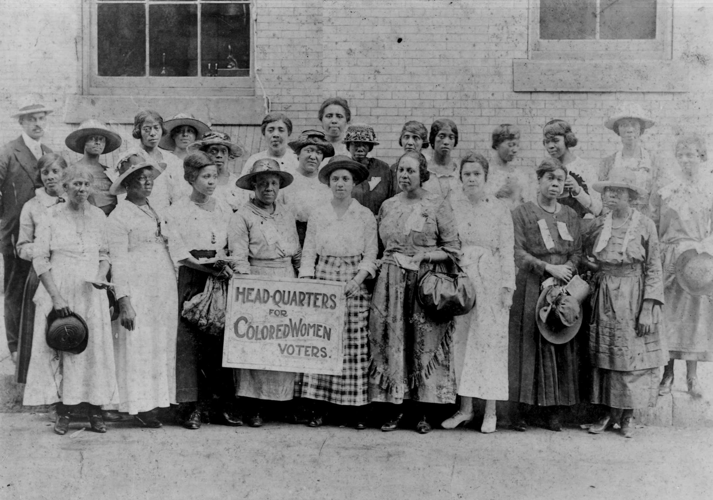 Black and white photograph of a group mostly woman standing in front of a building. They are holding hats and one central figure holds a banner with the name of the headquarters.