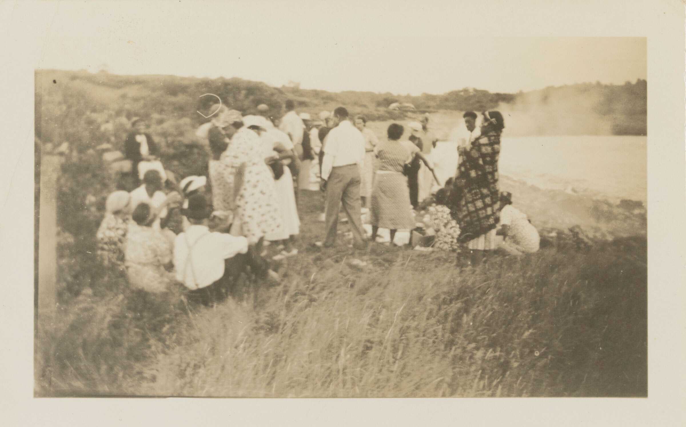 A black-and-white photograph of a group of people socializing on a grassy hillside. A large group of twenty-three men and women are sitting and standing with their backs to the viewer.