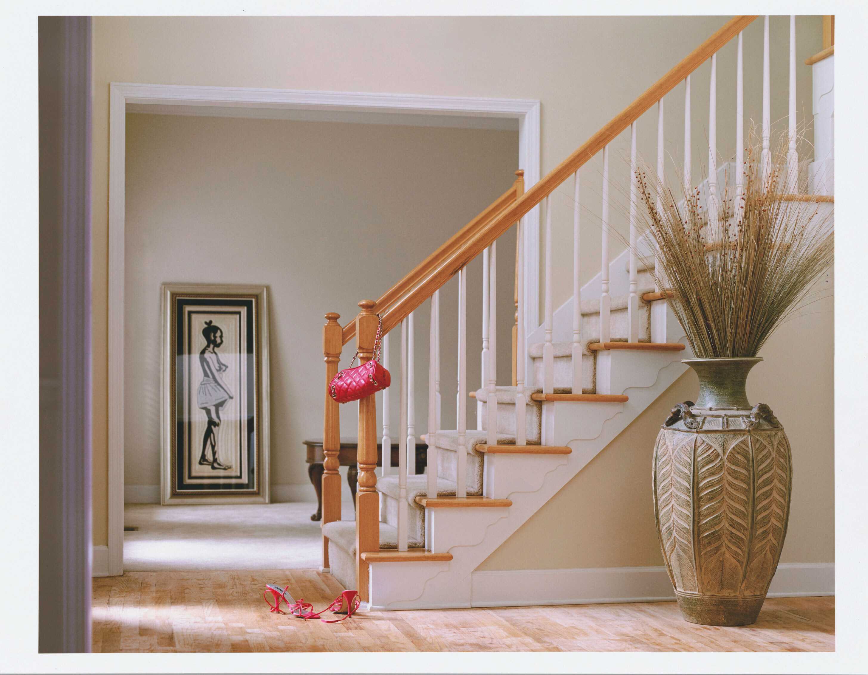 Color photograph showing the entryway to a home, showing a stairwell with a pink purse hanging from banister and a pair of hot pink high-heeled shoes at the ase.  There is a large vase with tall grasses next to the stairwell and a large framed picture of a woman, leaning against the wall in the distance..