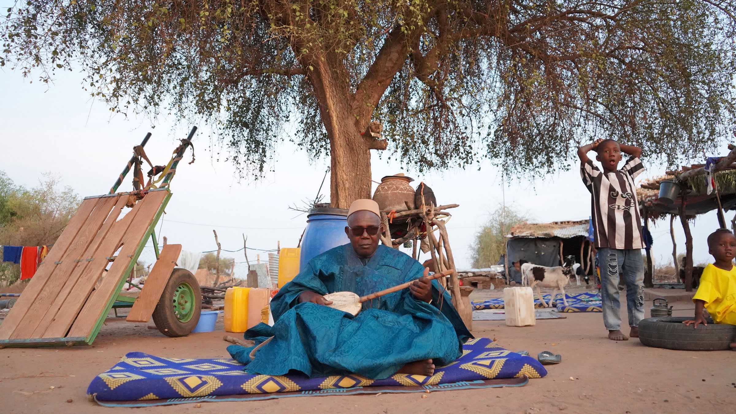 A griot, a West African storyteller, sits on the ground while playing a kora.