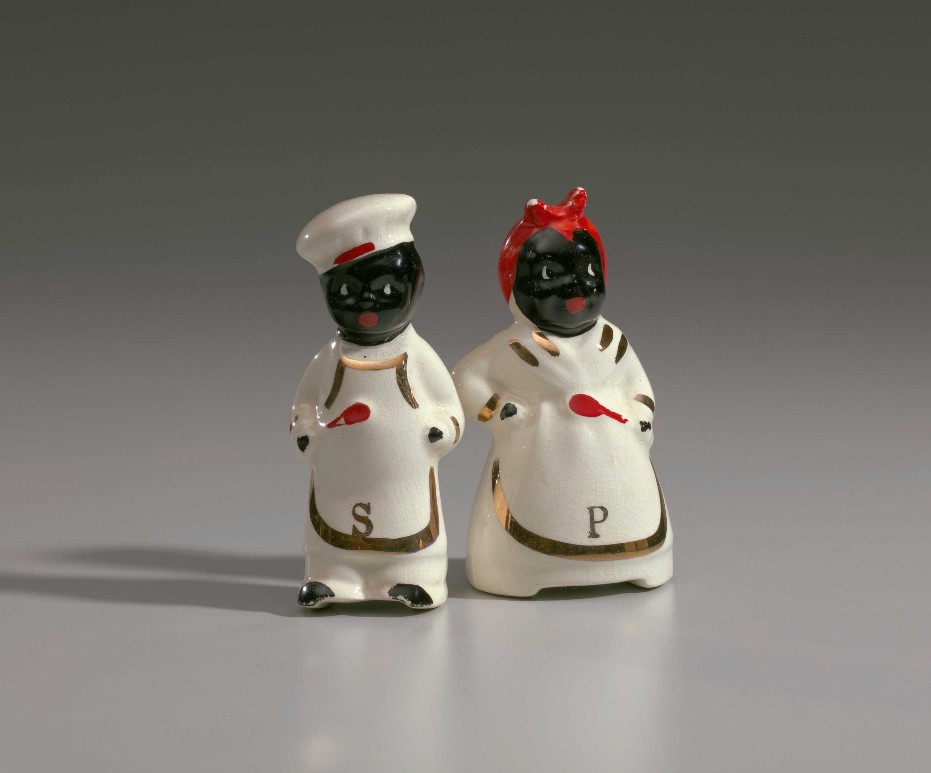 Ceramic salt shaker in the form of "Chef," who is depicted wearing a white chef's uniform, an apron outlined in gold, black shoes, and a white chef's hat with a red mark on the hat rim. "Chef" is holding a red spoon with his proper right hand. Ceramic pepper shaker , in the form of "Mammy," who is depicted wearing a white dress, white apron edged in gold and a white scarf with gold decoration and a red kerchief covering her head. “Mammy” is holding red spoon with her proper left hand. There is a gold [P] painted on the bottom center edge of the apron.