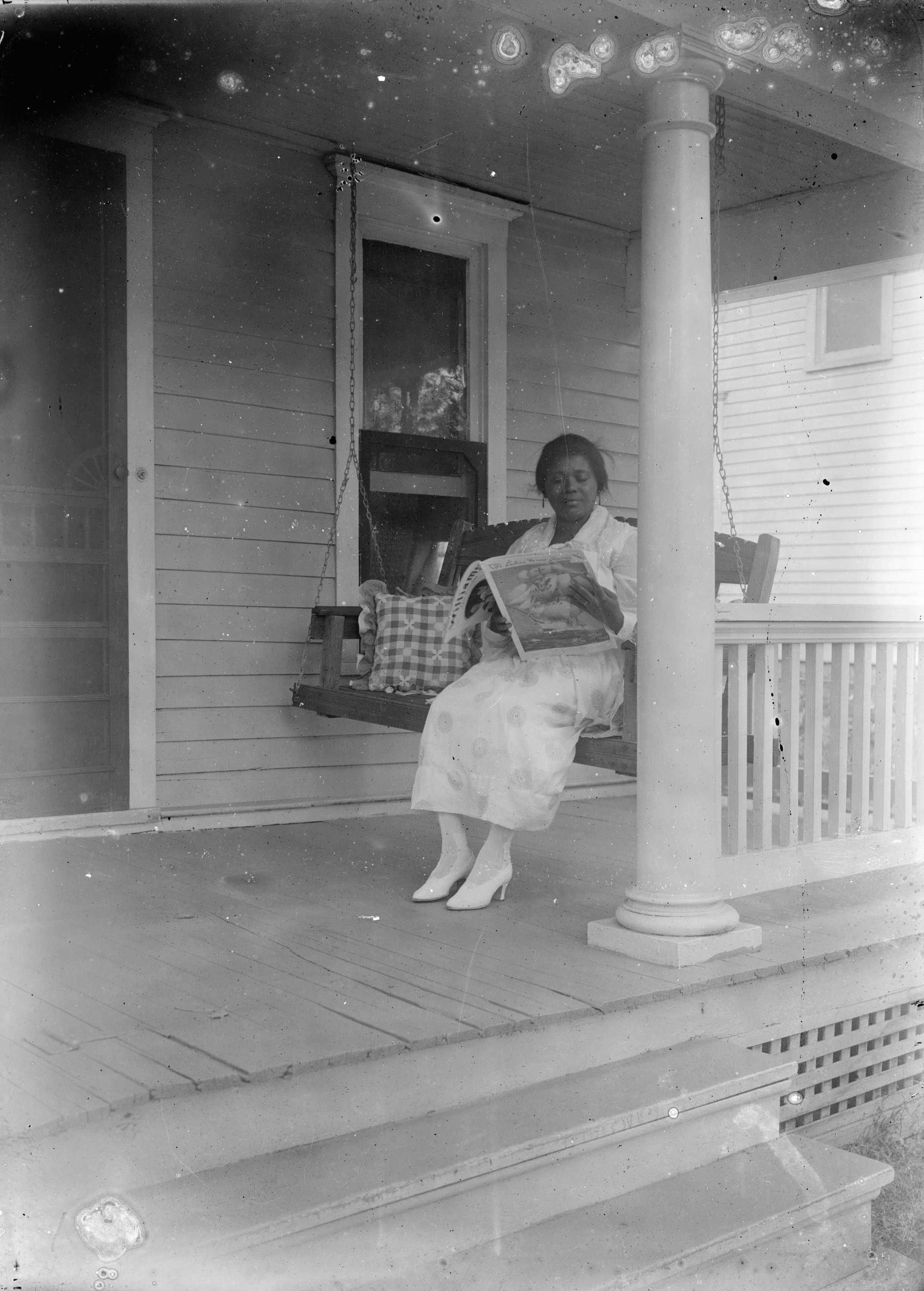 Woman sitting on a porch swing wearing a white dress and white hight heeled shoes.  She is reading a copy of Ladies Home Journal